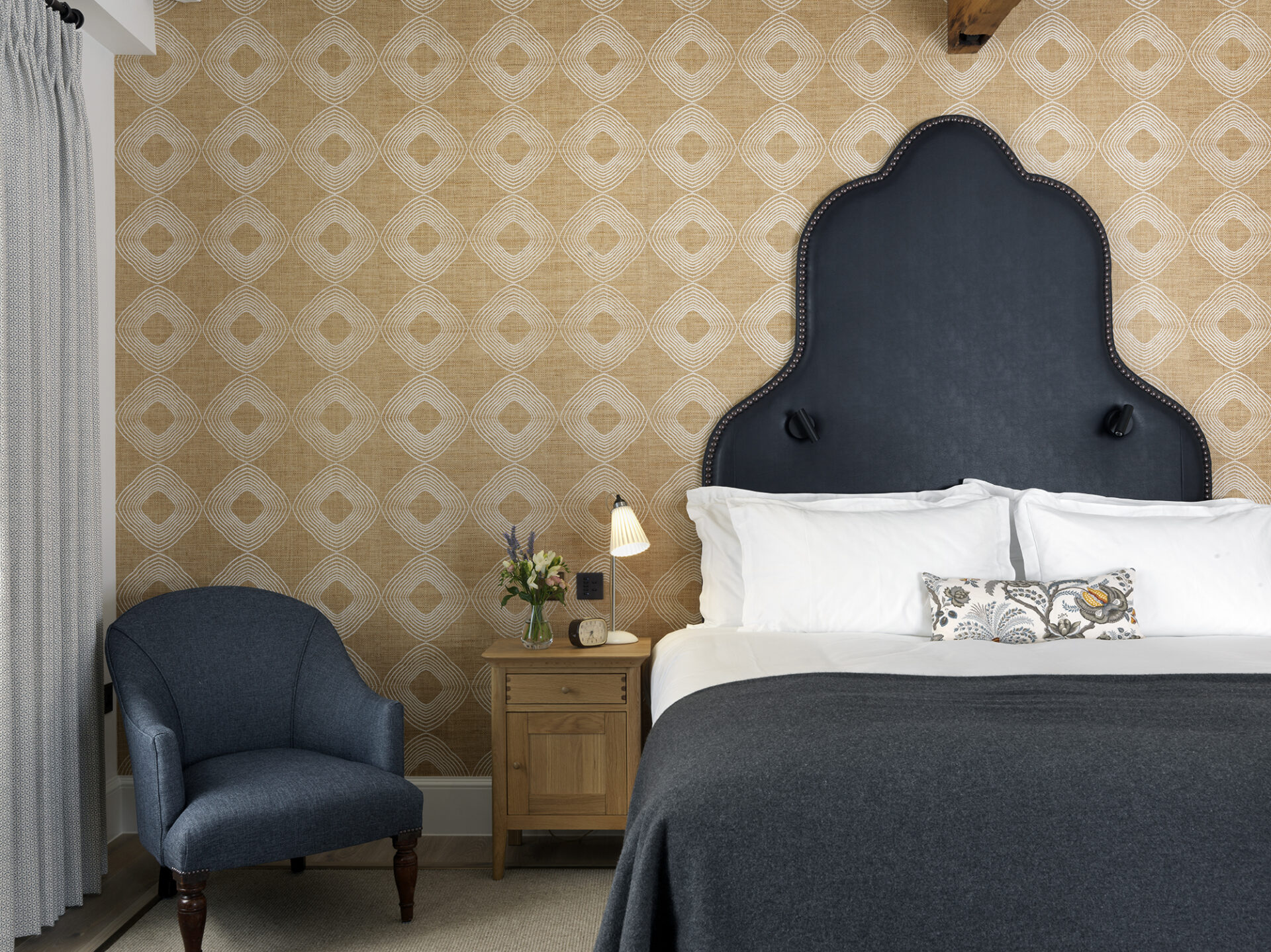 A cozy bedroom reminiscent of luxury five-star hotel suites in London features a dark upholstered headboard bed with white linens and a decorative pillow. A wooden nightstand with a lamp and flower vase sits beside it, while a blue armchair graces the corner against geometric-patterned wallpaper.