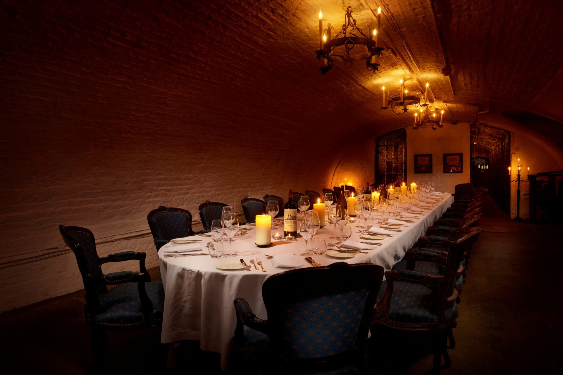 A long, elegantly set dining table with candles is surrounded by vintage chairs in a dimly lit, arched brick room at The Stafford London, with chandeliers hanging from the ceiling.