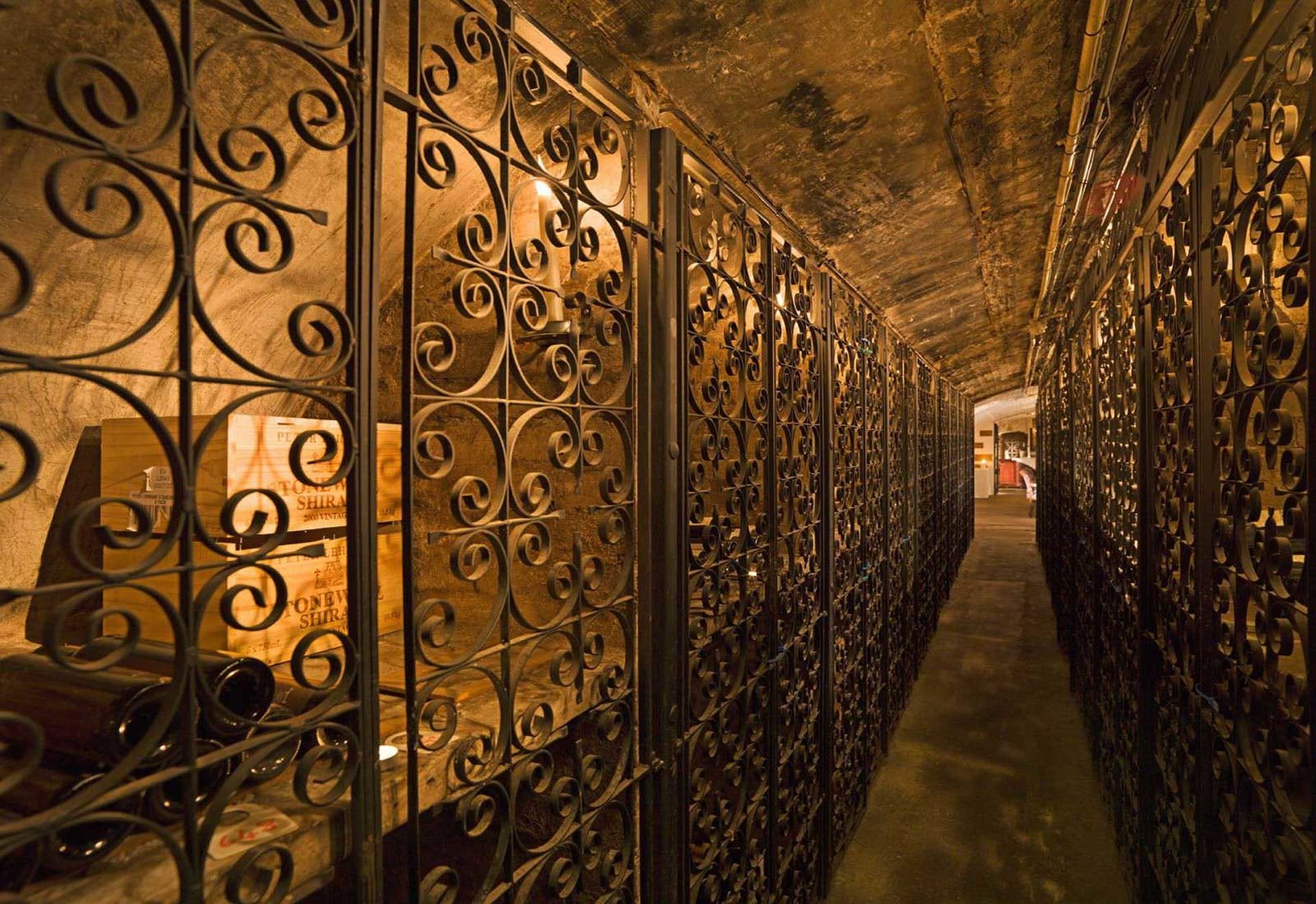 A dimly lit wine cellar with ornate wrought iron gates lining both sides, revealing wine bottles and wooden crates stored inside the locked compartments—an inviting setting for an intimate wine tasting along the narrow corridor.