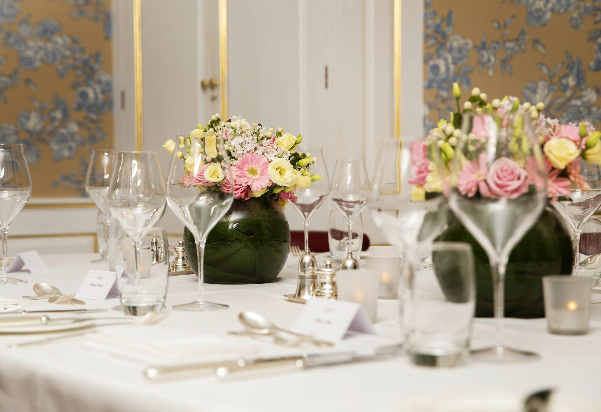Elegant table setting with clear wine glasses, silver cutlery, white name cards, and round green vases of pink and yellow flowers. Soft lighting and floral wallpaper create a refined atmosphere for unforgettable private dining events.