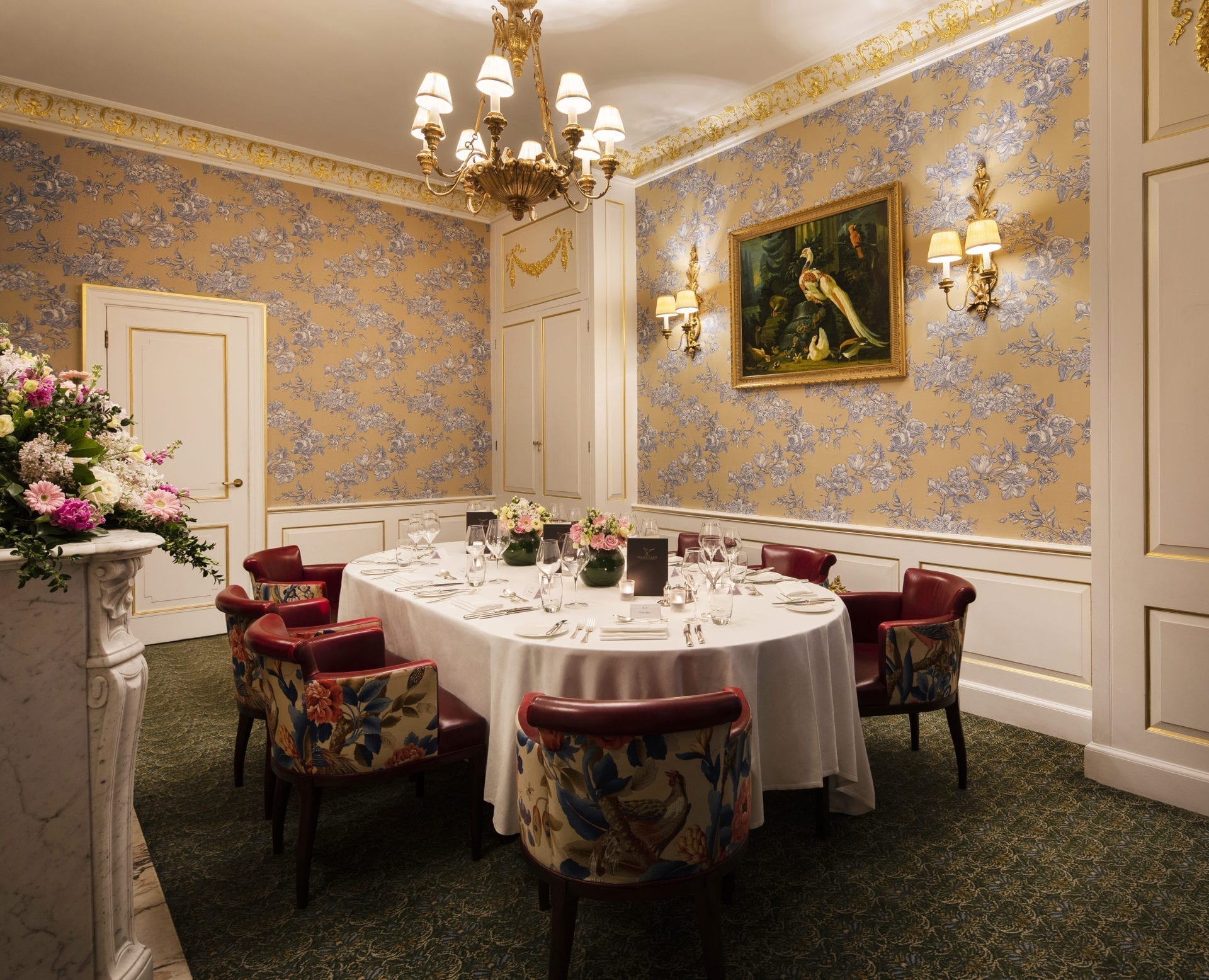 Elegant dining room with oval table set for eight, red chairs with floral patterns, ornate wallpaper, chandelier, wall sconces, flower arrangements, and a painting of birds. The refined setting is perfect for classic gatherings or intimate meetings.