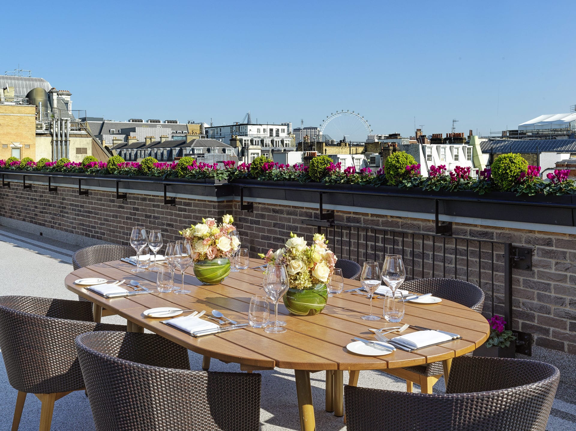 A rooftop dining area in a Luxury Five-star Hotel Suite London features a round wooden table set for six, surrounded by woven chairs and flower arrangements. City buildings and a large Ferris wheel shine under the clear blue sky.