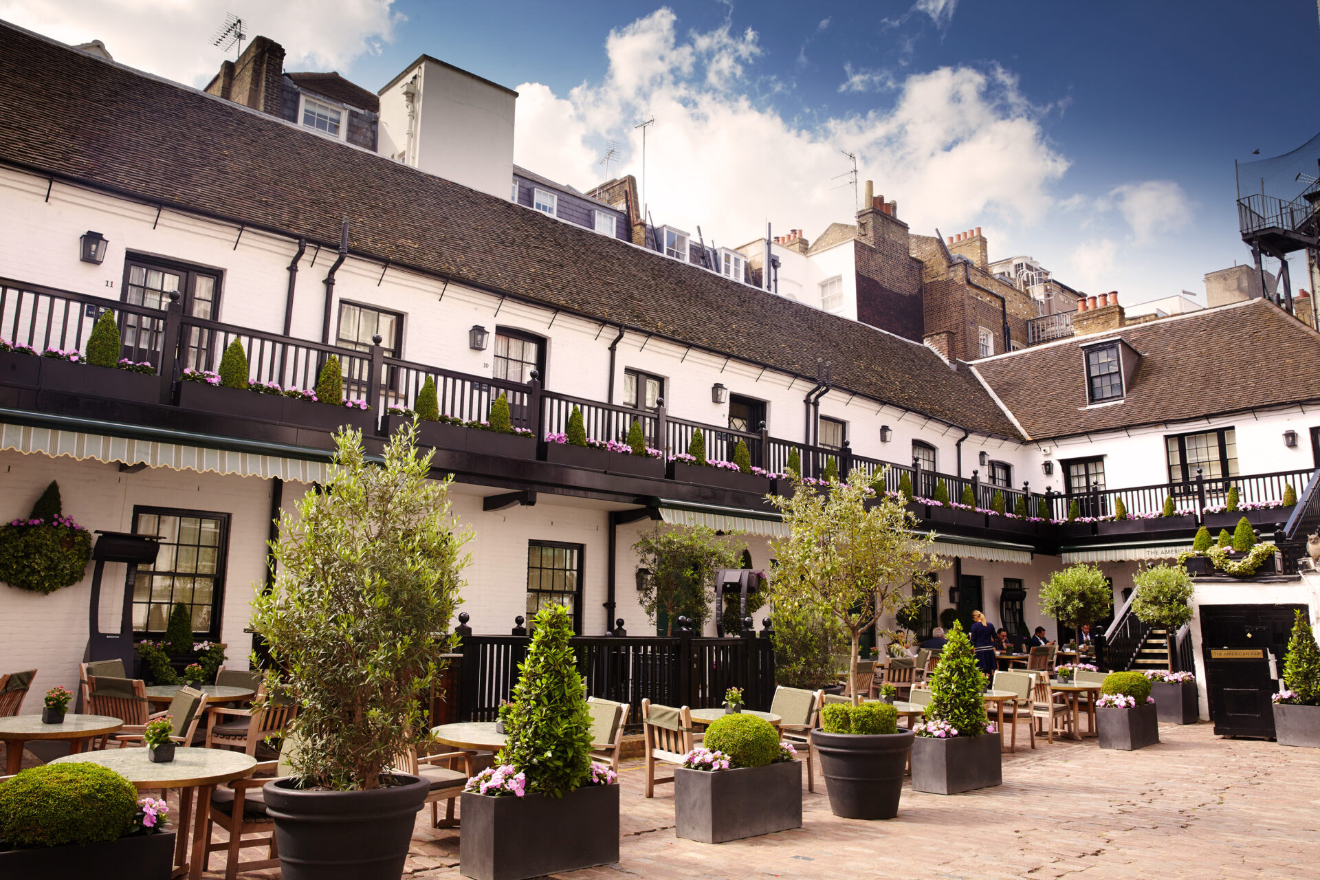 Sunny courtyard at The Stafford London with outdoor tables and chairs, potted plants, and a white two-story building with black trim and balcony. People relax under a blue sky dotted with clouds, enjoying the inviting space.