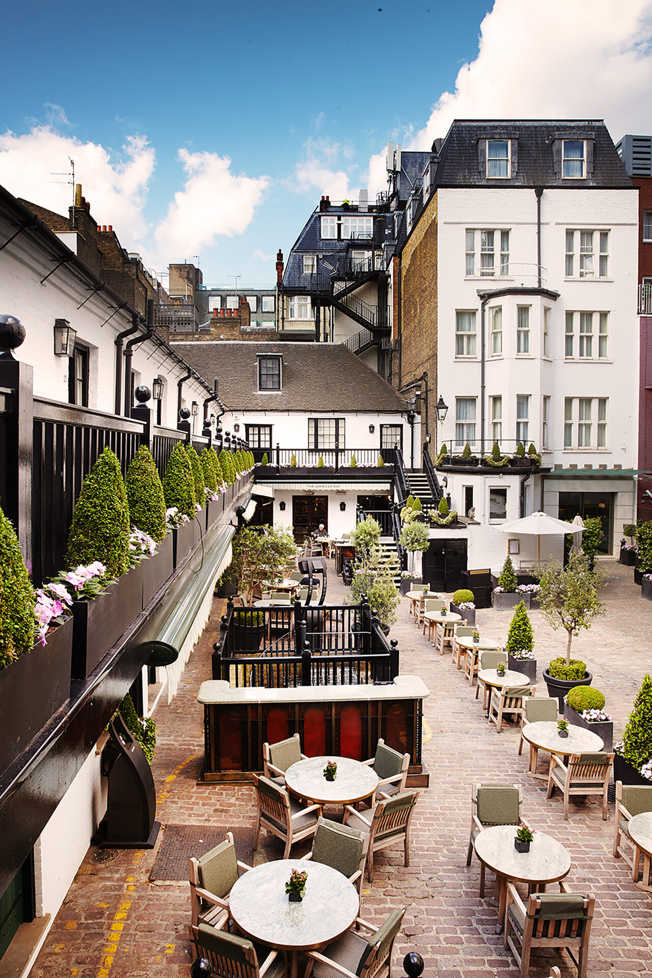 A charming outdoor courtyard at The Stafford London with round tables and wooden chairs, surrounded by potted plants and greenery, set against white buildings with balconies and black railings—perfect for dining or socializing.