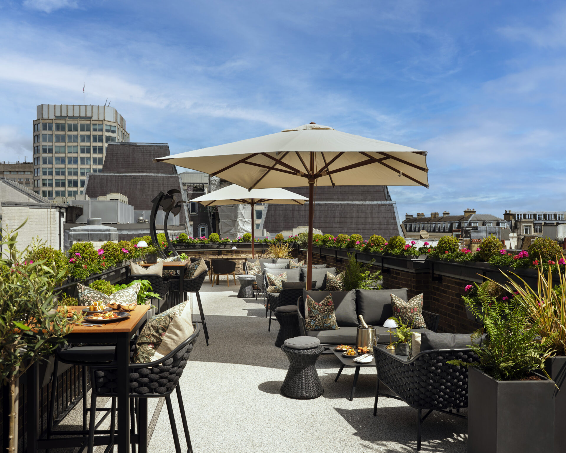 A stylish rooftop terrace with cushioned seats, tables set for private dining events featuring food and drinks, large umbrellas, potted plants, and city buildings in the background under a blue sky.