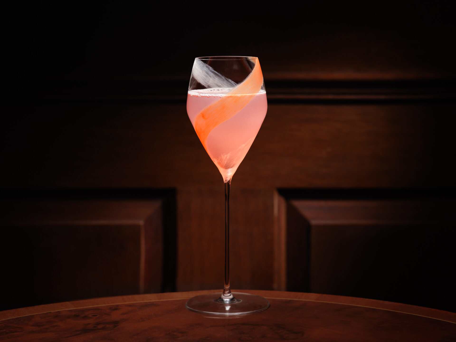 A pink cocktail in a stemmed glass sits on a wooden table against a dark background. The drink is garnished with an orange twist spiraling inside the glass.