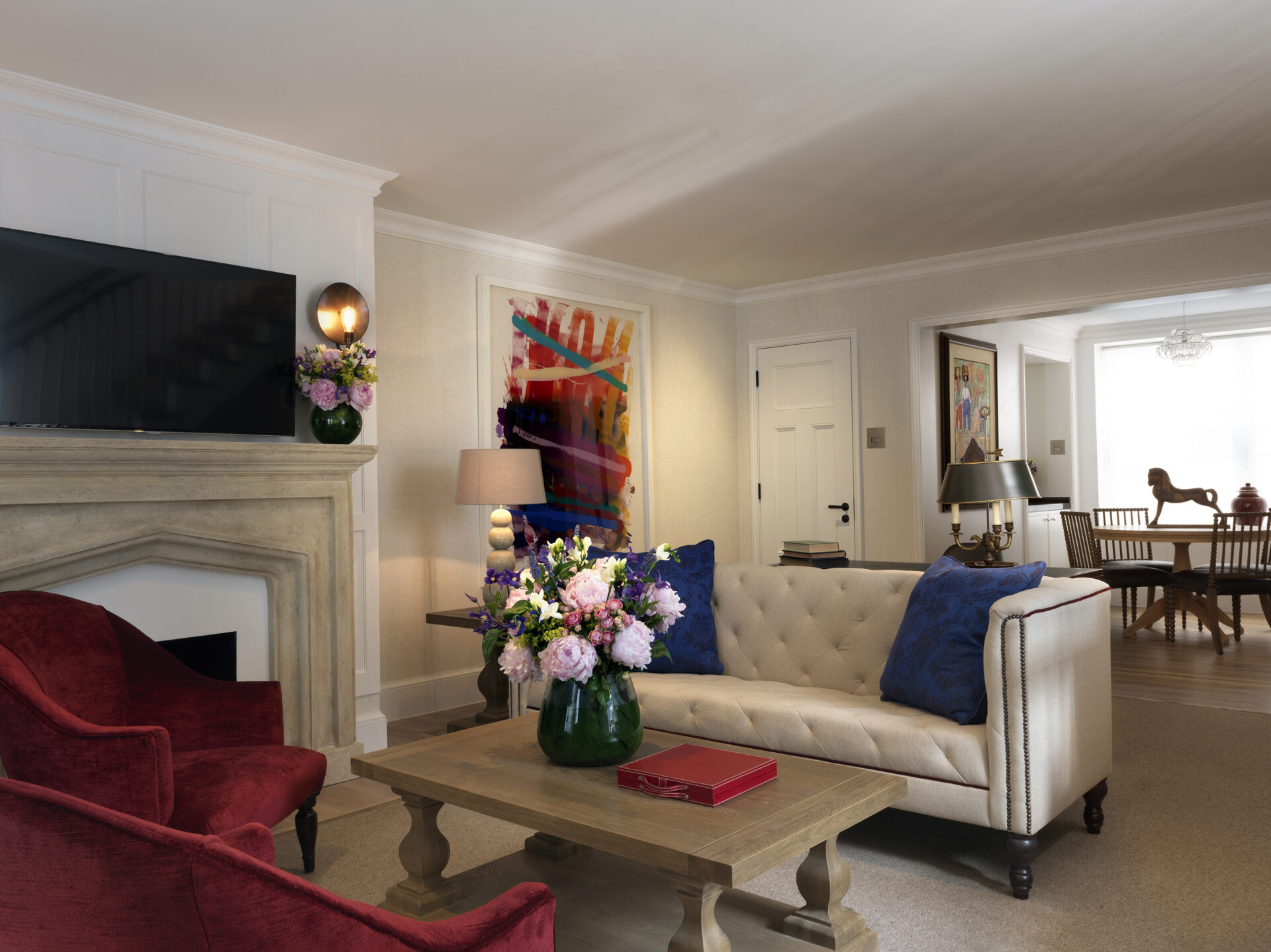 An elegant living room features a beige tufted sofa and two red armchairs around a wooden coffee table, where a vase of flowers stands. To the left, a fireplace rests below the TV. A vibrant abstract painting adds flair, much like the offers at The Stafford London in the dining room beyond.