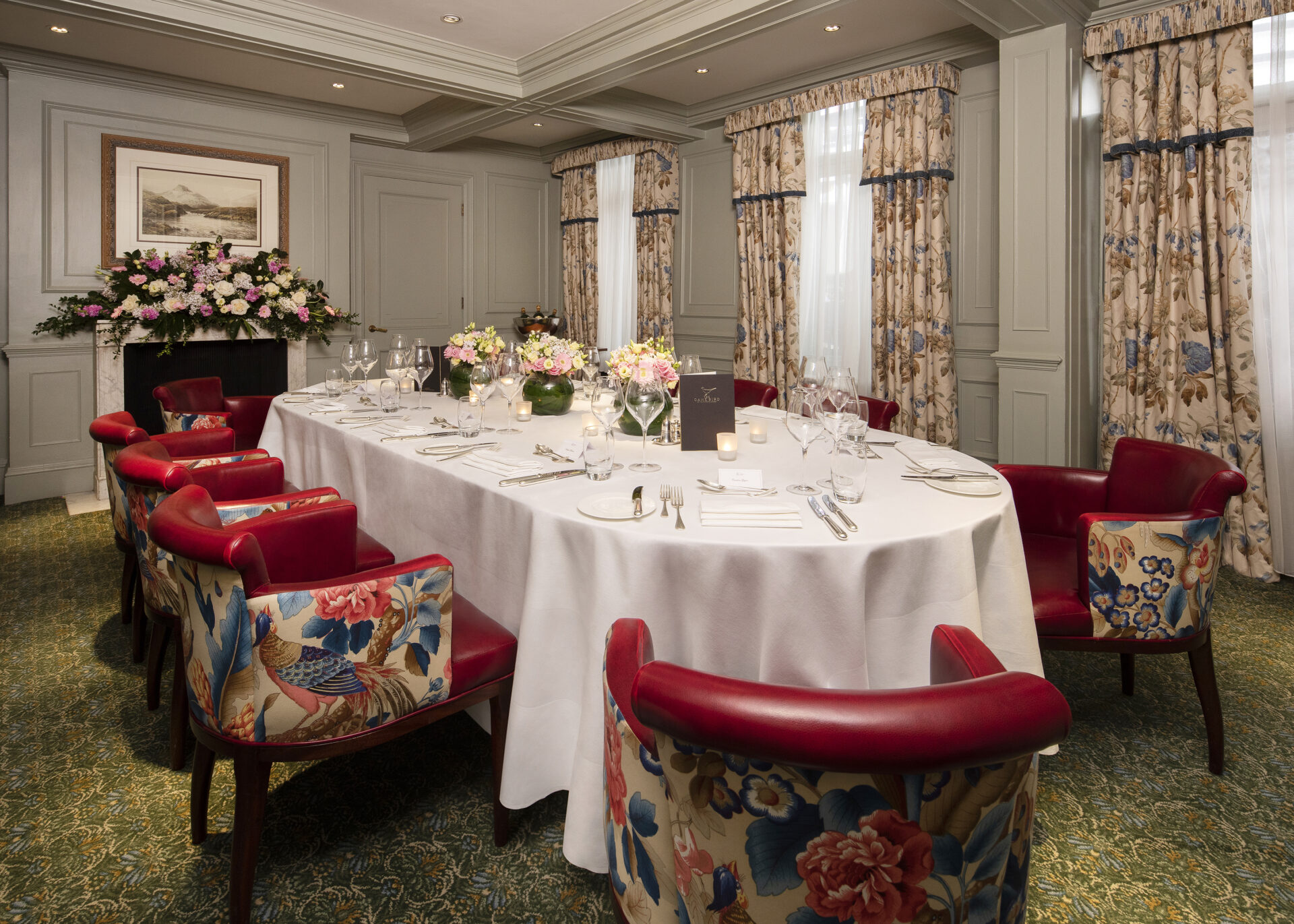 A formal dining room features a long table covered with a white tablecloth, set for eight with fine china and floral centerpieces. The room has patterned floral curtains, red and floral upholstered chairs, and paneled walls with a framed painting.