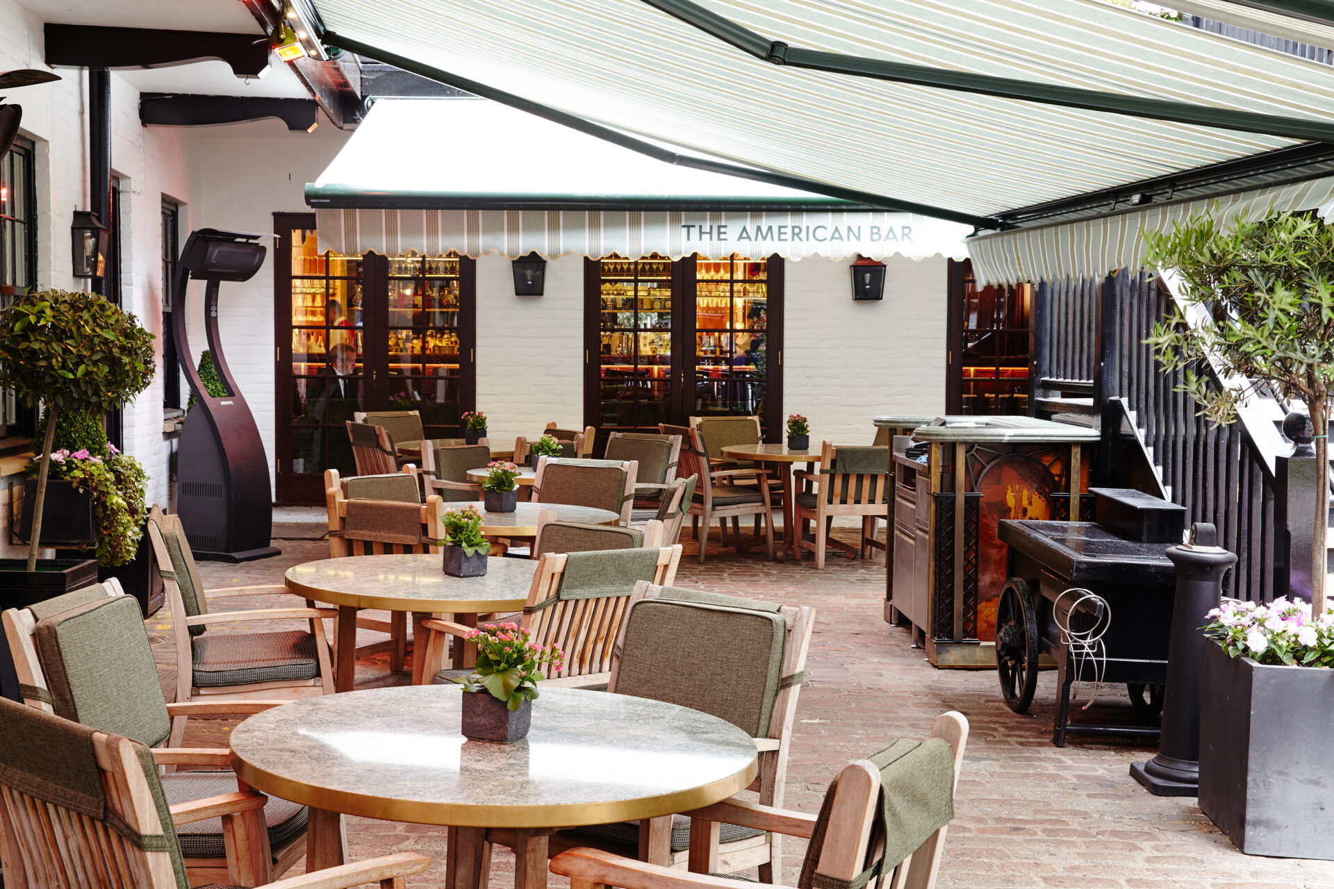 The outdoor patio of The American Bar features round wooden tables and green cushioned chairs under a shading awning. Potted plants add charm, while a vintage-style cart greets you near the entrance. For inquiries, a Contact Us sign ensures you can easily reach out with any questions.