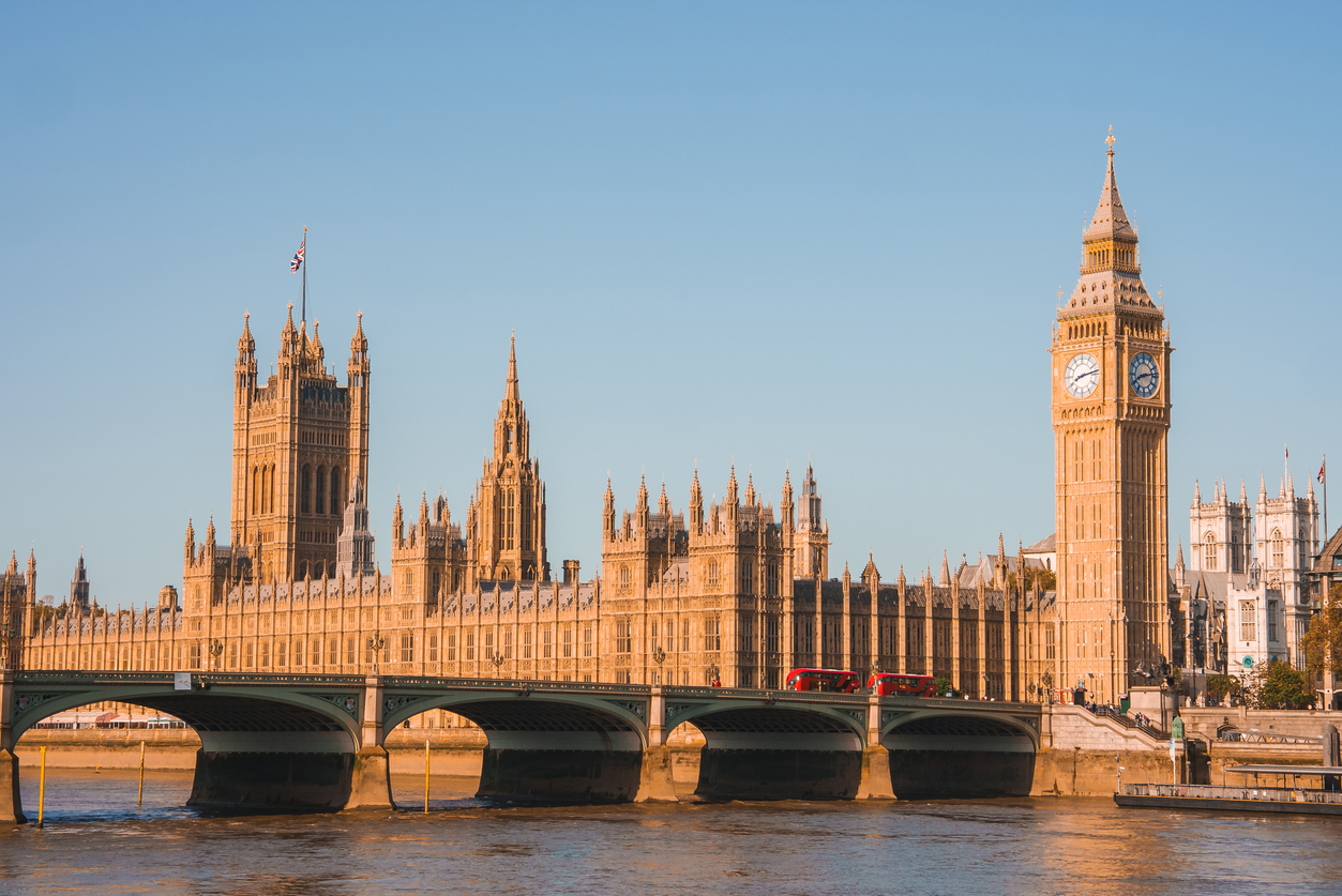 The image captures one of the historic attractions in London: the Palace of Westminster, with Big Ben standing tall. Bathed in warm sunlight, the scene features the River Thames in the foreground, where a bridge and red buses cross gracefully.