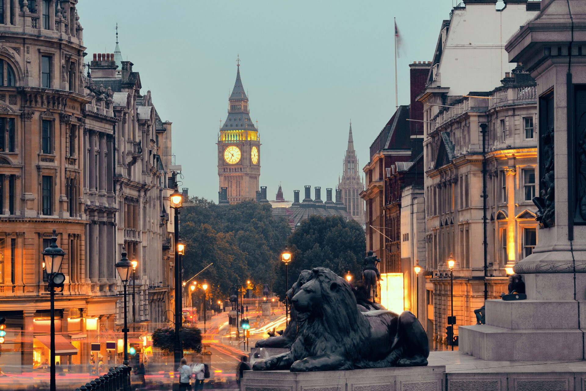 View of Londons Trafalgar Square with the Big Ben clock tower in the background showcases historic attractions. Lined with elegant buildings and a prominent lion statue, the scene captures a blend of city lights and motion.