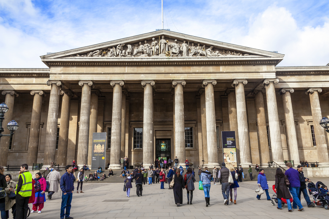 People gather outside a grand neoclassical building with large columns, likely a museum or historic site, in the heart of London. On a clear day in Mayfair, visitors and tourists of various ages walk and sit, enjoying the atmosphere before indulging in nearby shopping delights.