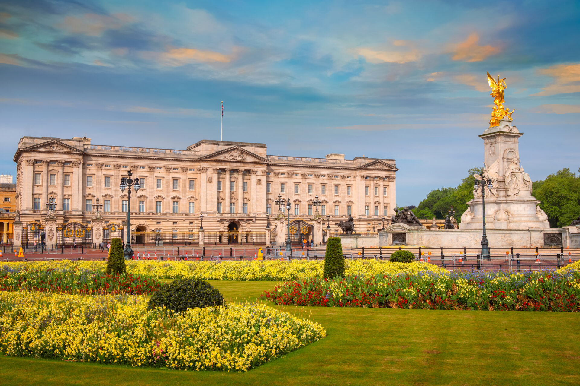 Buckingham Palace, one of Londons premier royal attractions, lies beneath a vibrant sky with colorful clouds. The foreground showcases manicured lawns and flowerbeds, while the Queen Victoria Memorial stands prominently with its striking golden statue.
