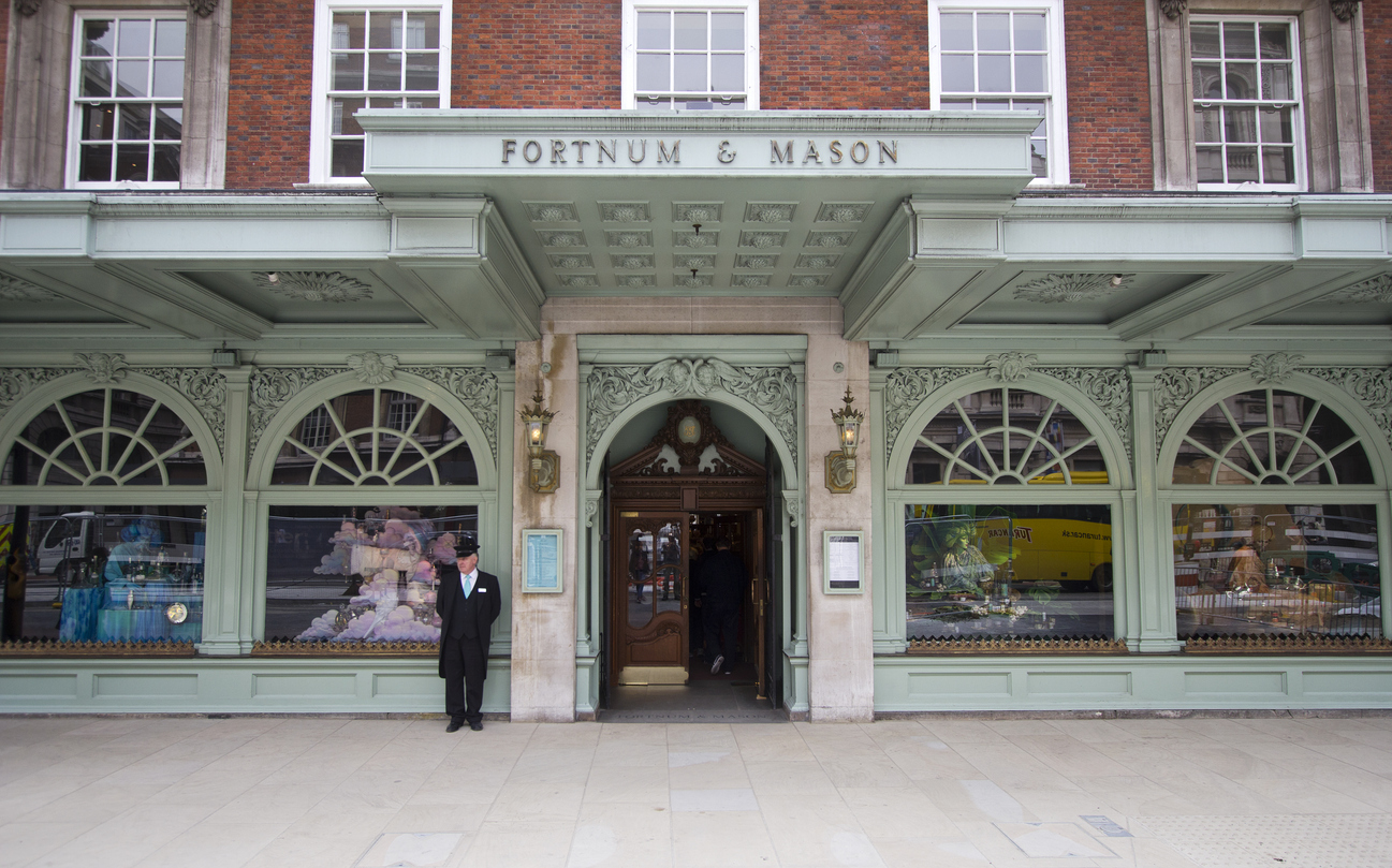 The exterior of Fortnum & Mason in Mayfair showcases a luxury department store with an ornate green facade. A suited doorman stands next to display windows brimming with colorful products, epitomizing London shopping. The stores name is prominently displayed above the entrance.