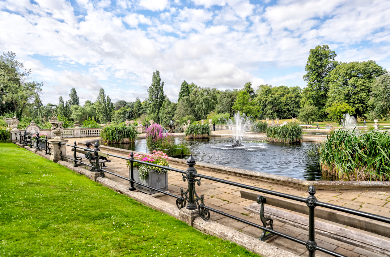 A royal attraction in London, this elegant park features a large rectangular pond with multiple fountains. Lush green grass, flowering plants, ornate stone urns, and a wrought iron fence surround the area, while trees and a partly cloudy sky complete the serene backdrop.