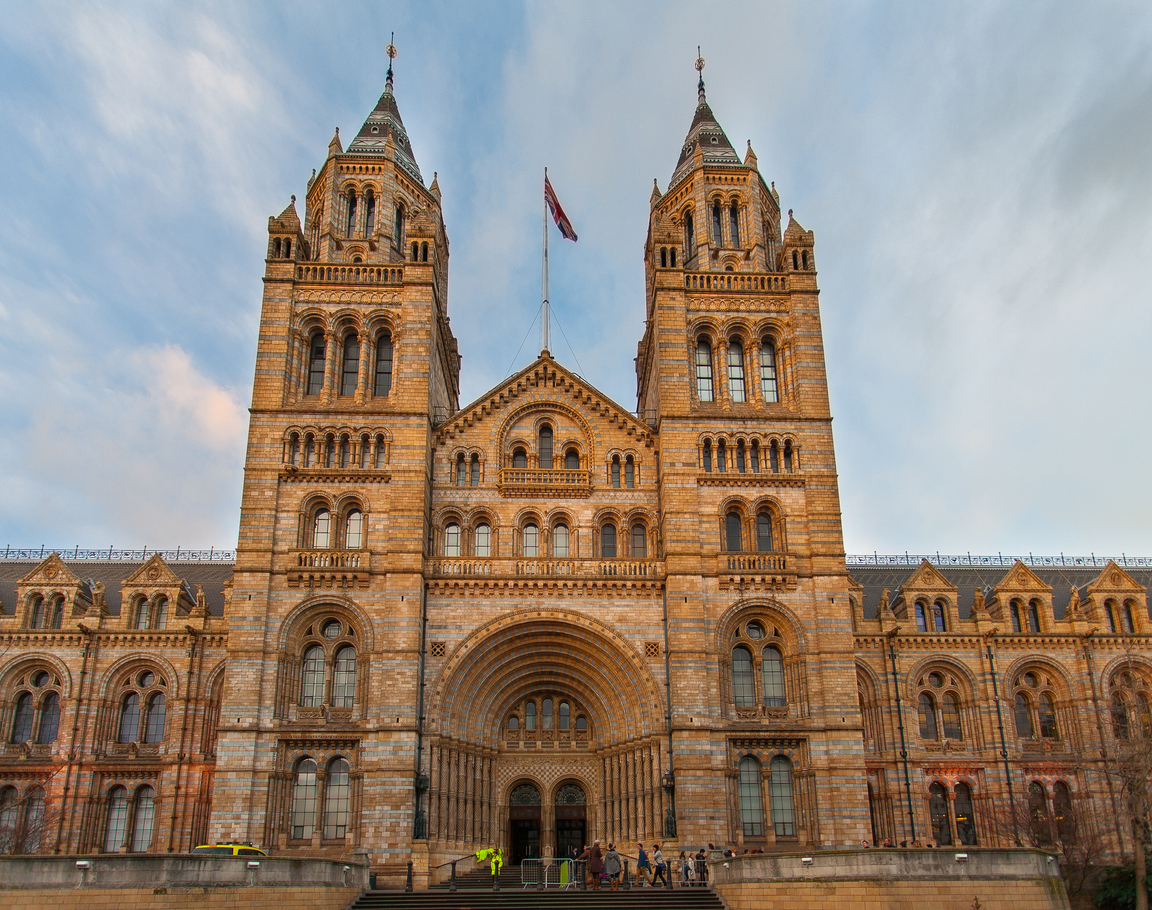 The image captures a large, historic building in Mayfair, London, featuring two prominent towers and intricate stonework. A flag flies atop the central section. Ornate arches and windows adorn the façade, while people gather at the entrance, possibly after a day of shopping.