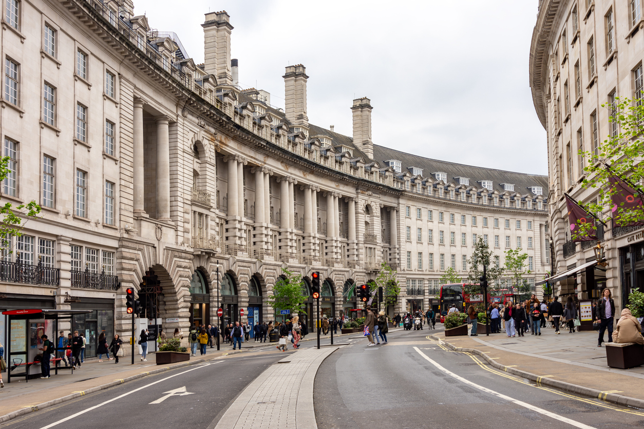 Street view of a curved road in Mayfair, lined with grand, historic buildings. Pedestrians stroll along the sidewalk, and red buses navigate the street. The architecture boasts large windows, columns, and stone detailing under a cloudy London sky.