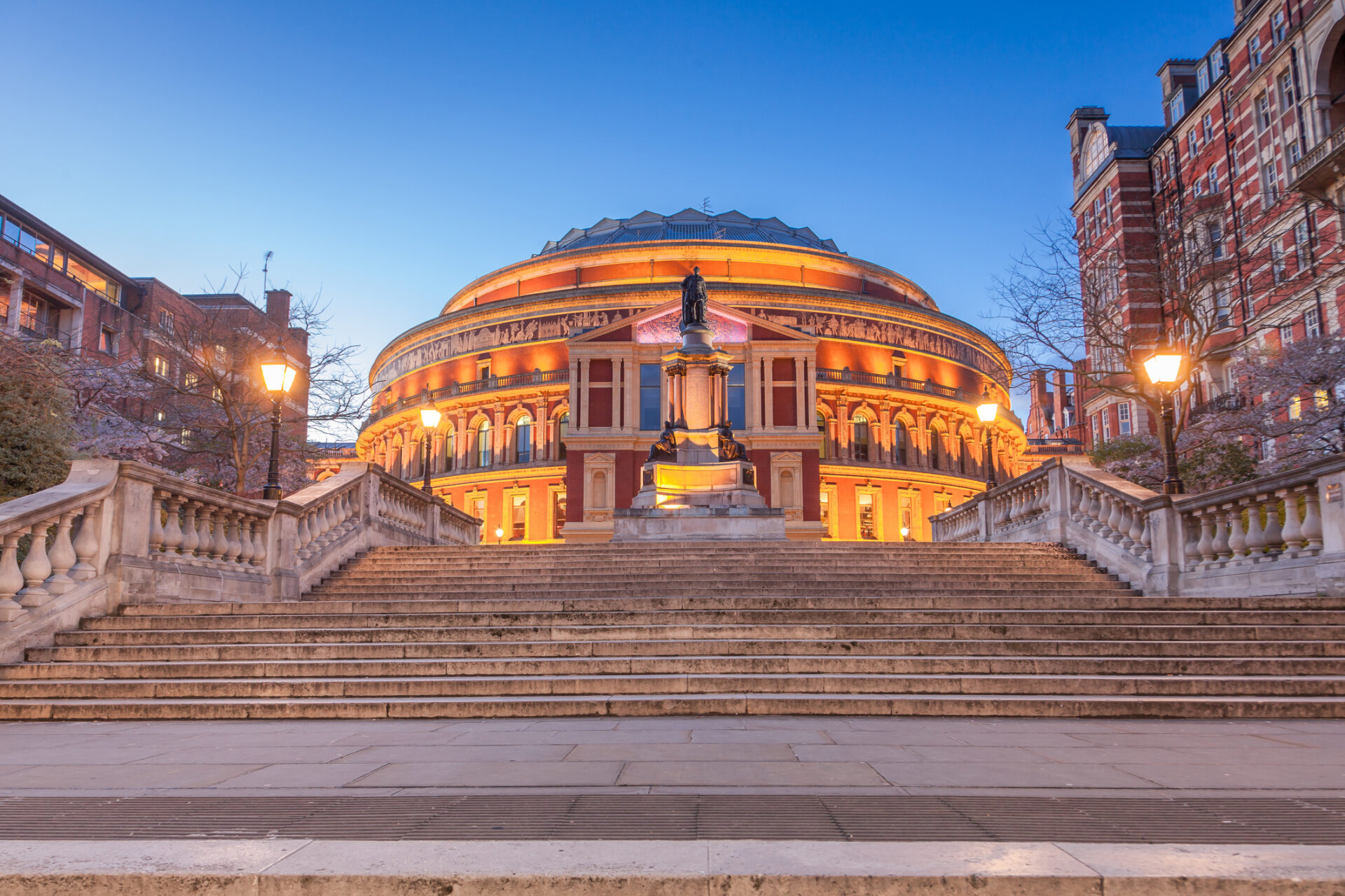 The image captures the Royal Albert Hall in London at dusk, its grand architecture illuminated splendidly. A statue adorns the steps leading up to the entrance, flanked by glowing street lamps. Not far away lies Mayfair, famed for its upscale shopping and vibrant atmosphere.