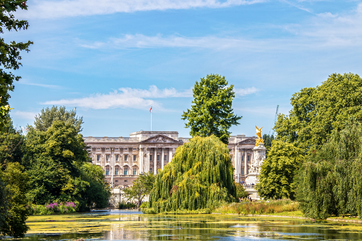 Buckingham Palace, one of the premier royal attractions in London, stands majestic with its scenic gardens in the foreground. Lush green trees and a tranquil lake reflect the serene setting under a blue sky, while a golden statue peeks into view on the right.