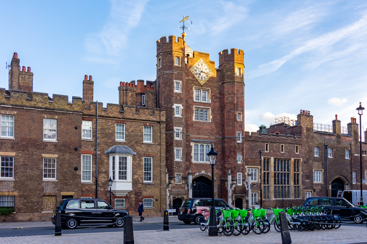 Street view of St. Jamess Palace in London, a royal attraction with its iconic red brick facade and clock tower. Black cabs are parked nearby, and a row of green rental bicycles awaits riders in the foreground under a clear blue sky.
