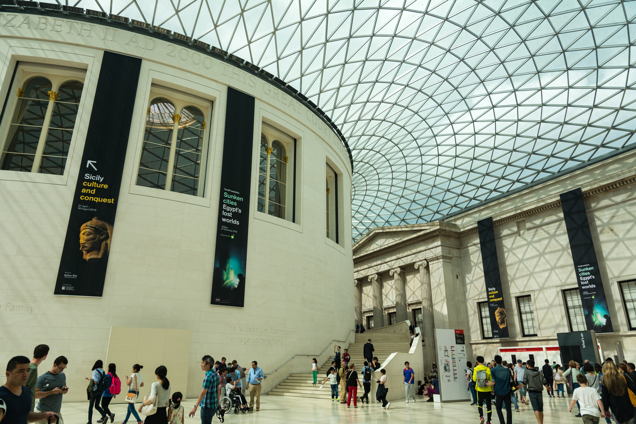 The spacious interior of a museum, reminiscent of Mayfairs elegant charm, features a large glass roof, stone walls, and vibrant exhibition banners. Visitors explore the bright and airy setting reminiscent of a London shopping experience while discovering captivating cultural displays.