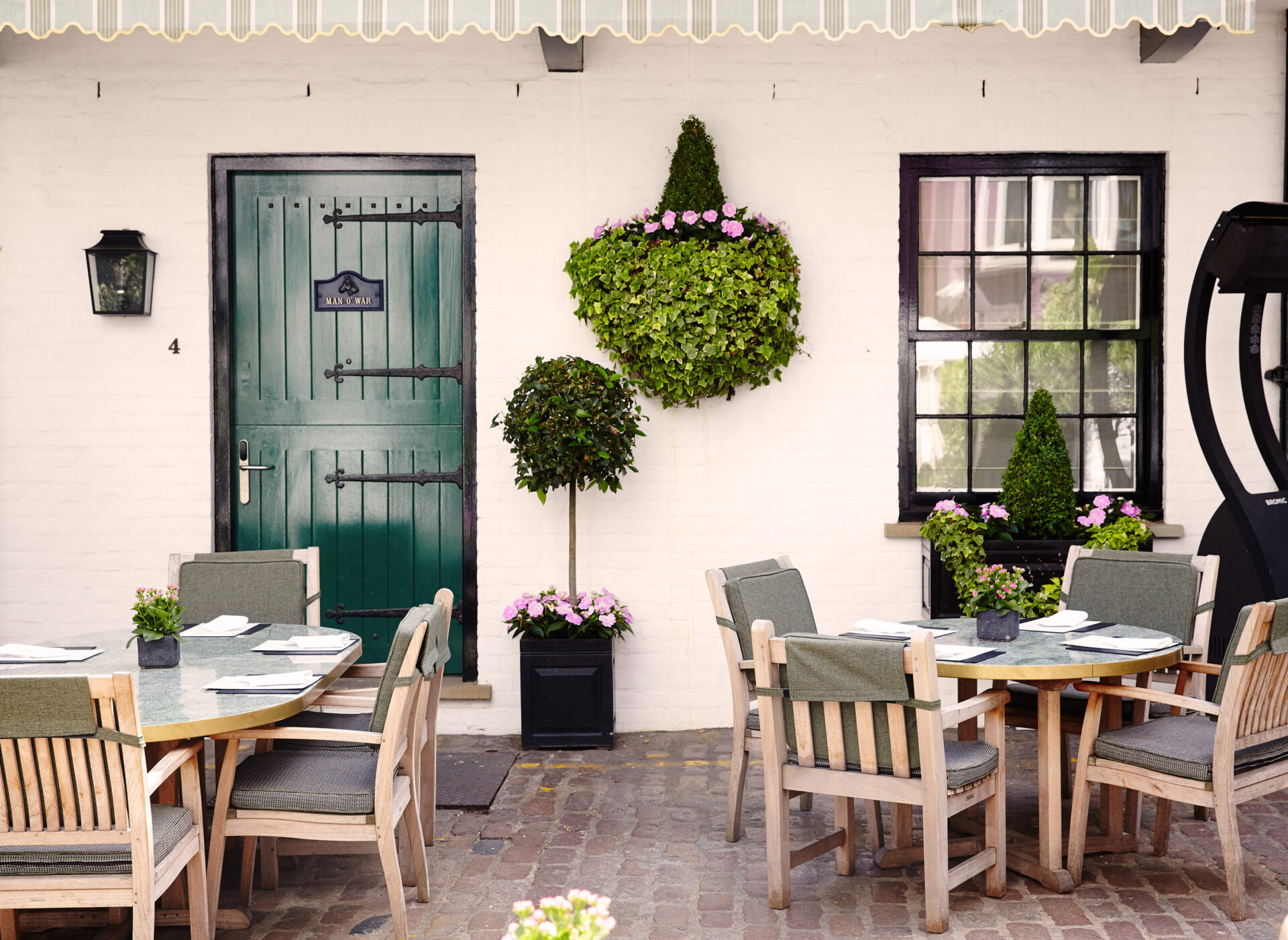 Step into this charming outdoor dining area featuring wooden tables and chairs on a cobblestone patio. A green door and windows accent the white brick wall, while news-worthy potted plants and flowers bring life to the space beneath a shading canopy.