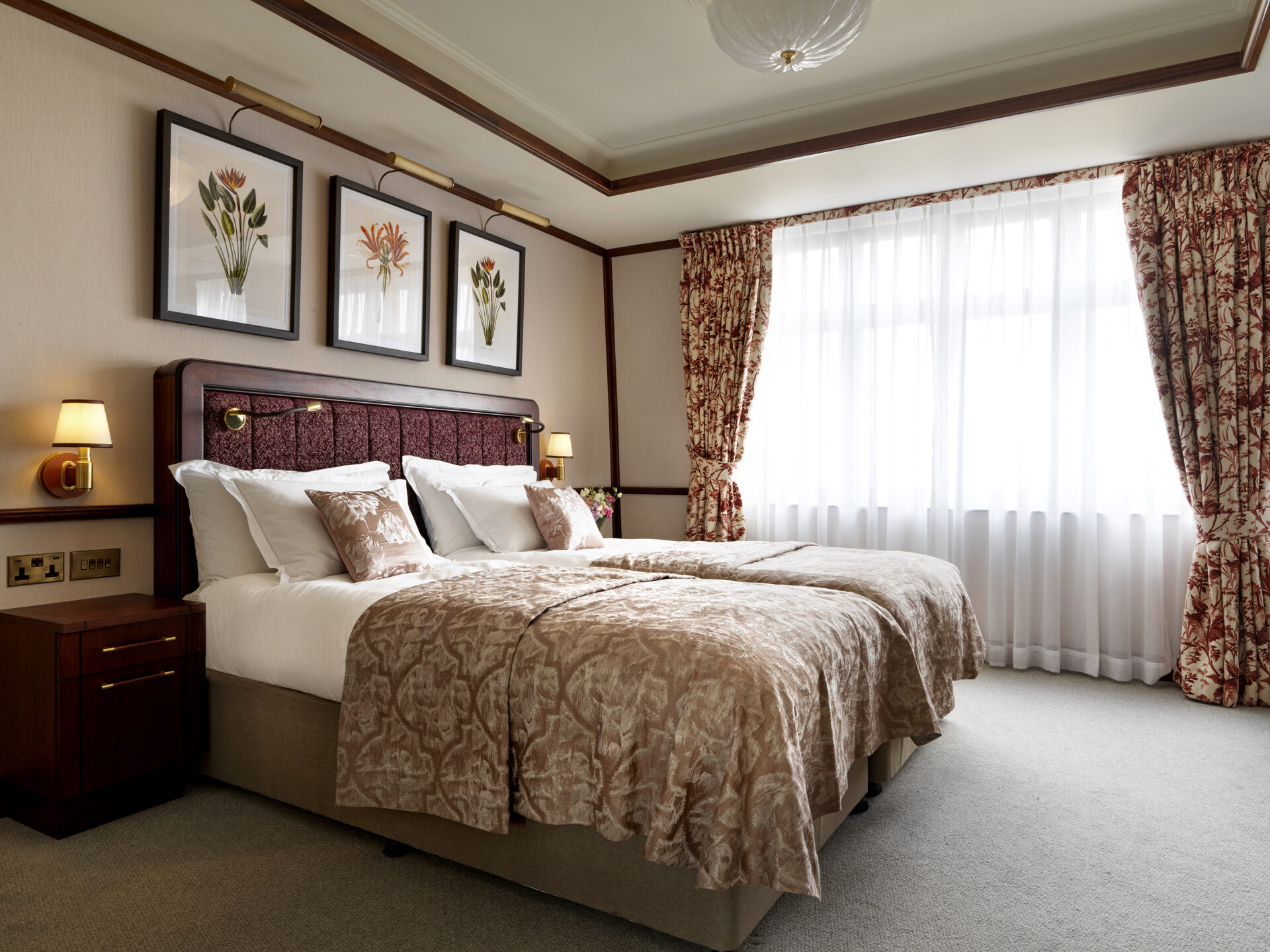 A hotel room perfect for a family getaway, with two neatly made twin beds featuring beige patterned bedding. The upholstered headboard is adorned with floral paintings above it. Two wall lamps and floral curtains frame a large window, creating a cozy atmosphere around the wooden nightstand.