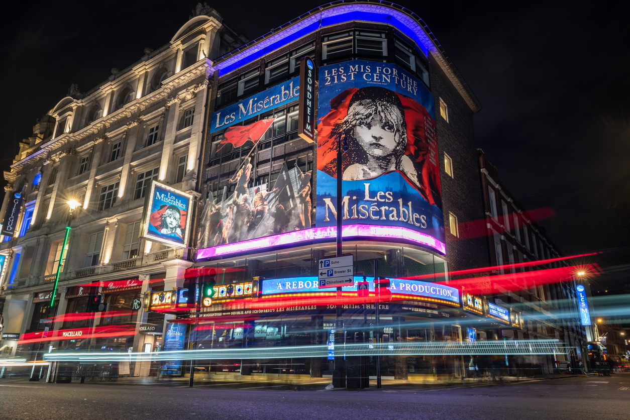 A city street at night in Mayfair features a brightly lit theater with large posters for Les Misérables on its facade. Light trails from passing vehicles streak across the foreground, adding dynamic movement to the scene, capturing the vibrant essence of London.