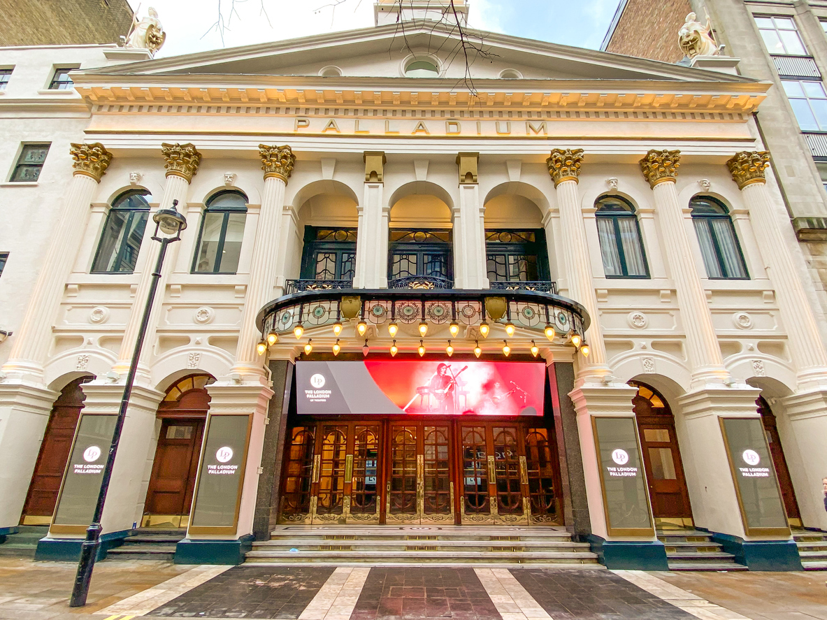 The exterior of the Palladium theater in Mayfair boasts tall columns and ornate decorations, blending seamlessly with the upscale urban setting. A digital marquee promotes upcoming events, inviting passersby to enjoy an experience as elegant as London shopping itself.
