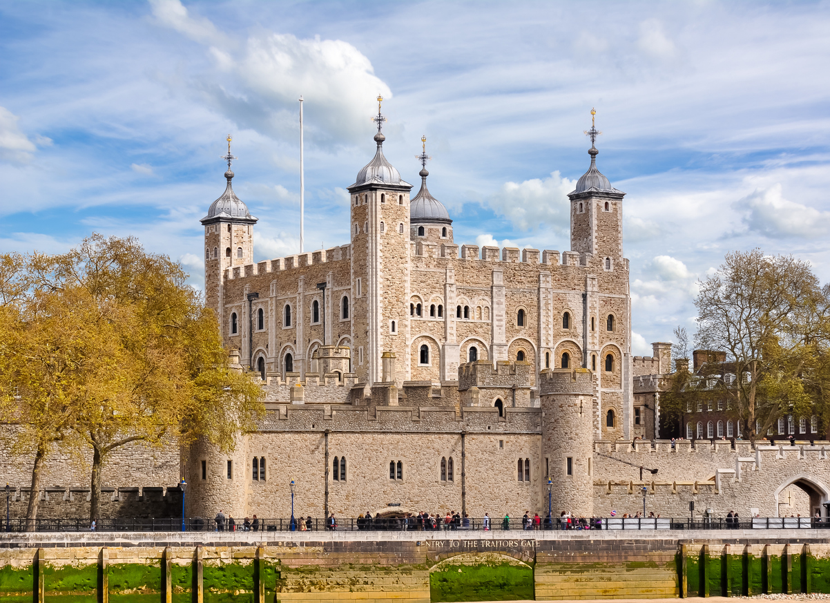 The image captures the Tower of London, one of the historic attractions in London, a stone fortress with multiple turrets under a partly cloudy sky. Green trees frame the scene as a line of people stroll near the entrance along the waters edge.