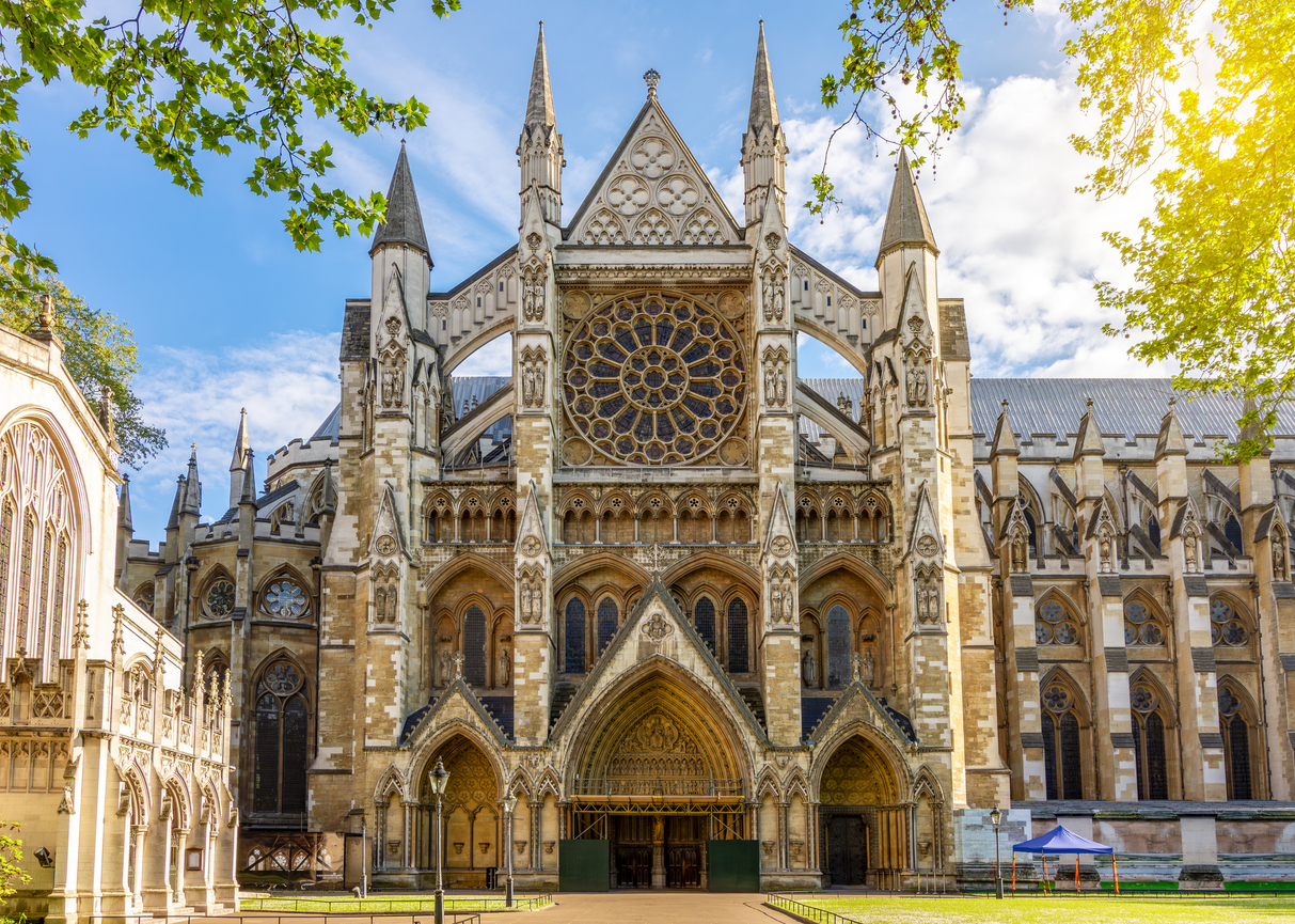 A grand Gothic cathedral facade, reminiscent of historic attractions in London, features intricate stonework and two large arched entranceways. Above them are decorative spires and a prominent circular stained glass window. The sun casts a warm glow, framed by lush green trees.