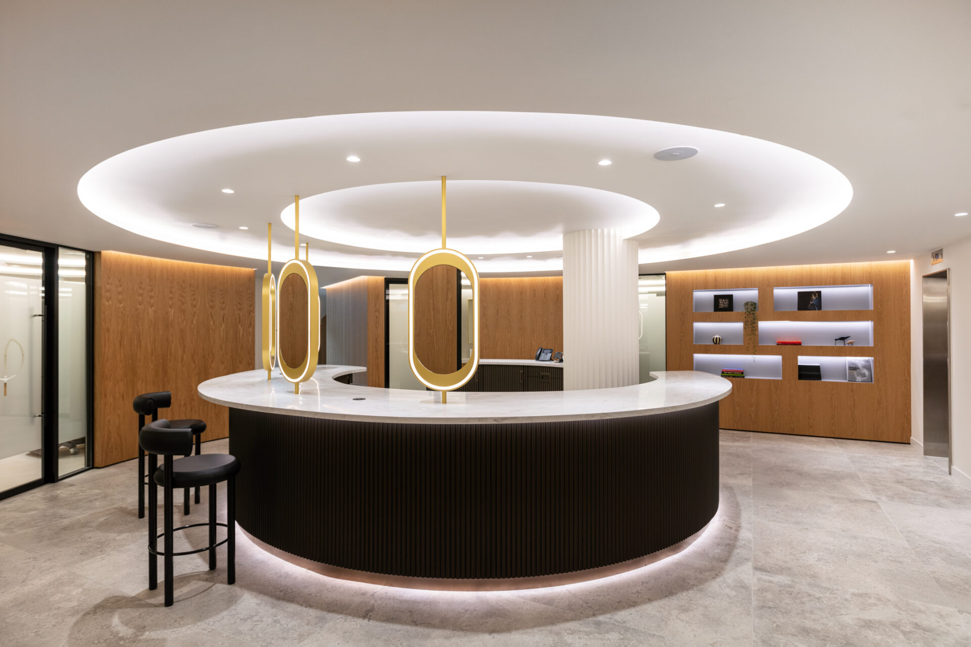 The modern reception area, reminiscent of Londons royal attractions, features a circular white counter, black barstools, and overhead circular lighting. Wooden panel walls with shelves display items against neutral-toned flooring and ambient lighting for a sleek, contemporary atmosphere.