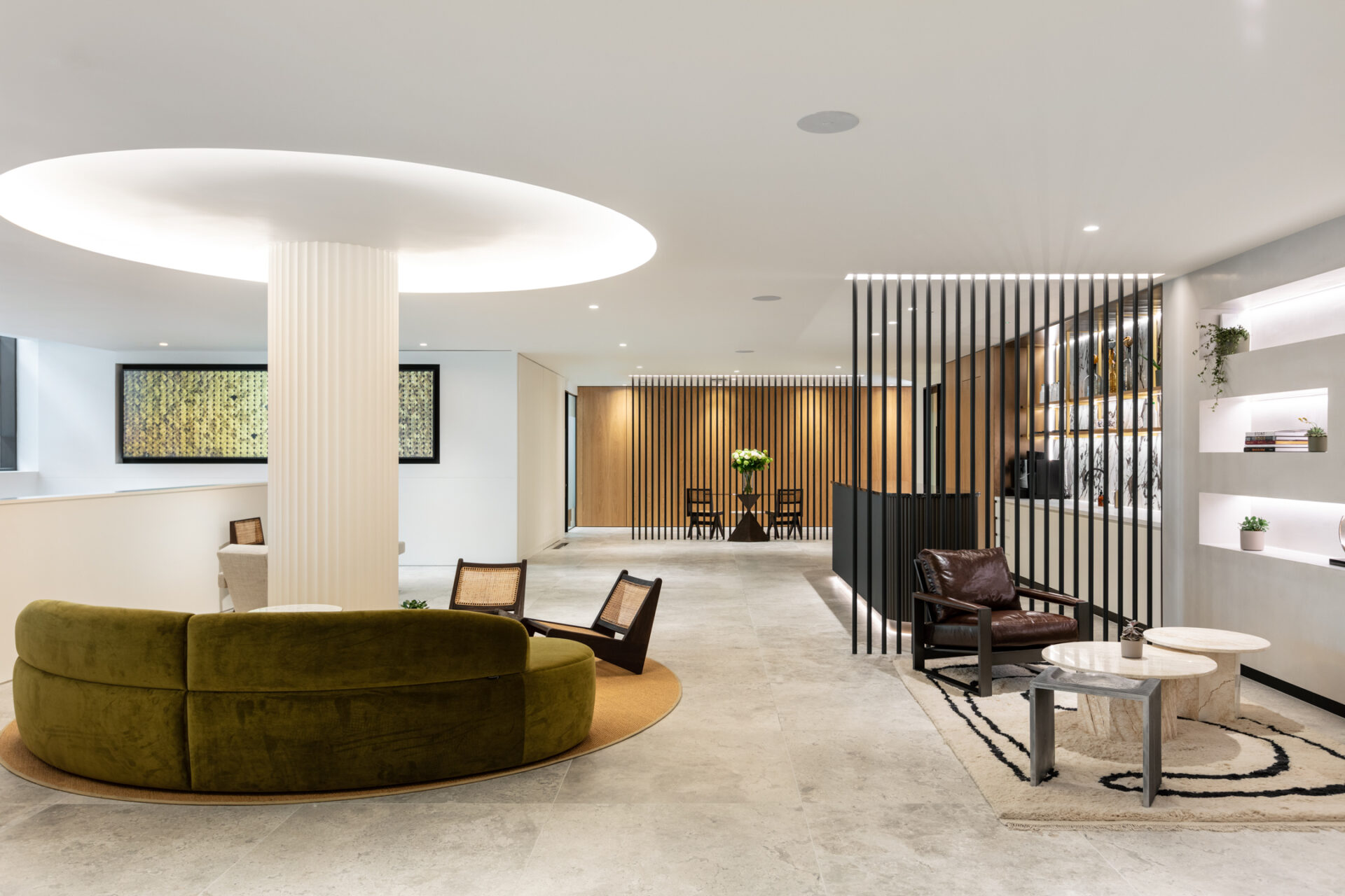 The modern lobby, reminiscent of royal attractions in London, features a circular olive-green sofa and wooden chairs around a small table. A round ceiling light illuminates abstract art, decorative shelving, and a sleek slatted partition leading to another neutral-toned room.