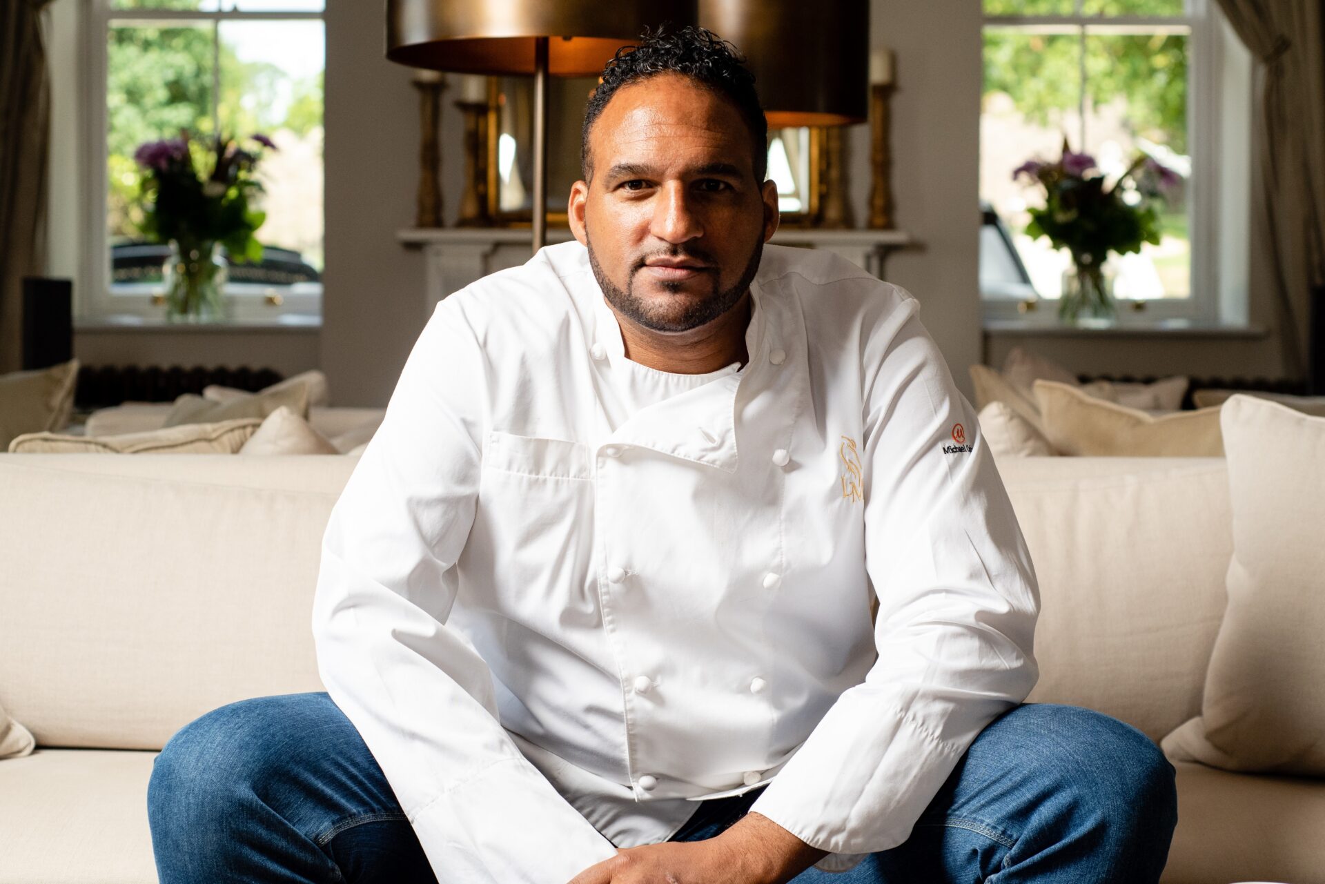 A man in a white chefs coat sits on a light-colored couch. He has short hair and a beard, and is looking directly at the camera. The background features floral arrangements and large windows.