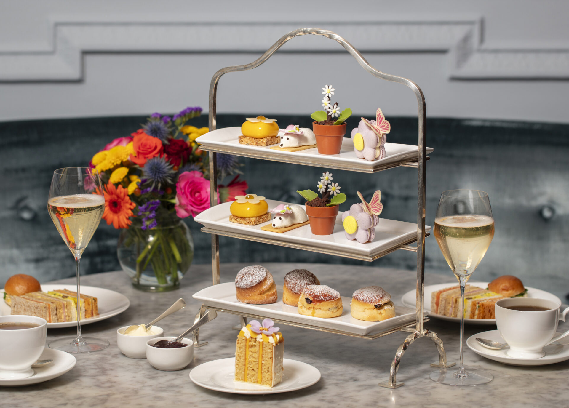 A three-tiered tray with assorted pastries, cakes, and desserts sits on a marble table alongside two glasses of sparkling wine—perfect for indulging in exclusive Dining Offers. A colorful bouquet and slices of layered cake complete the elegant setting.