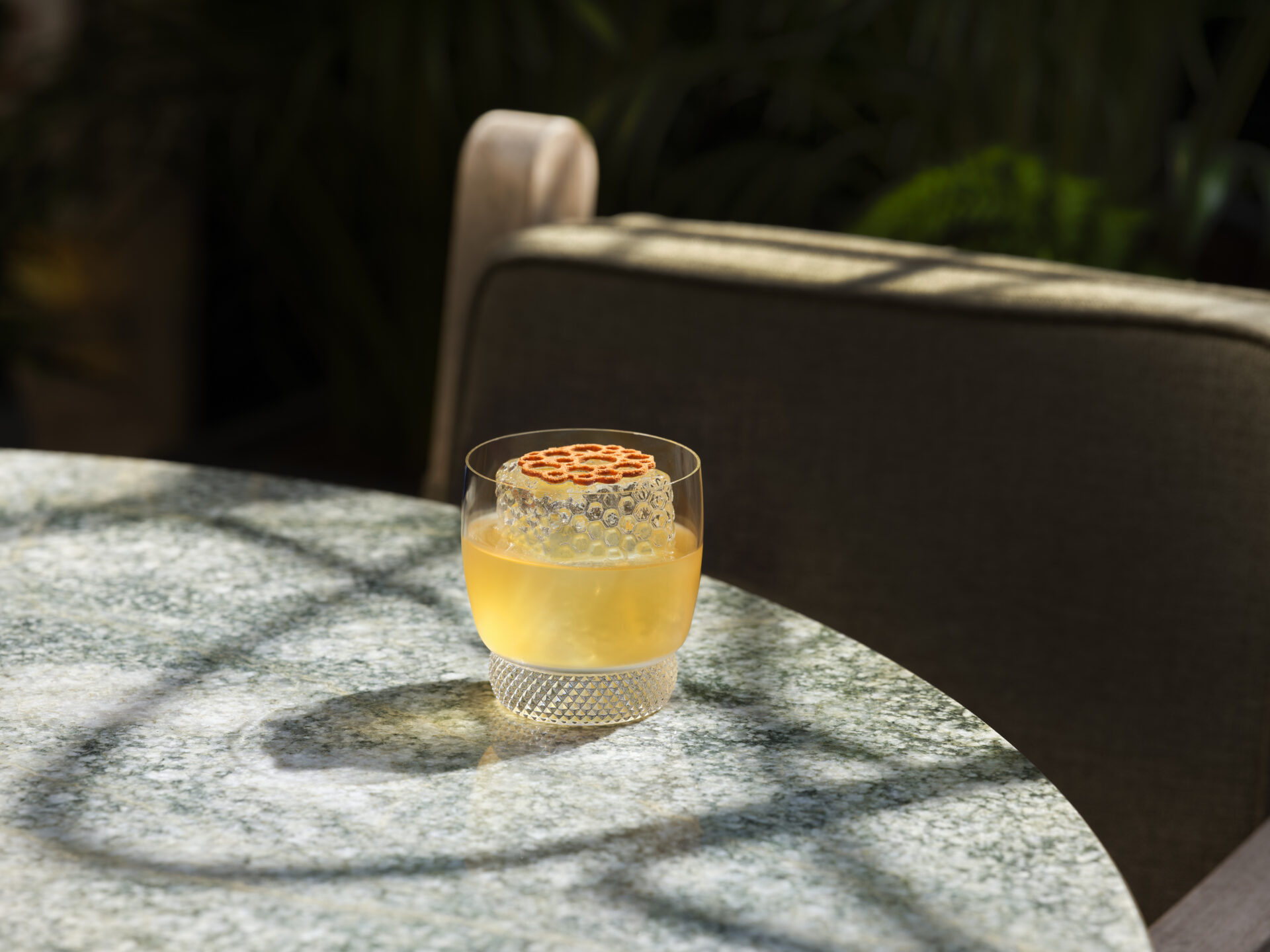A clear glass with a yellow-orange drink and decorative garnish sits on a round, marble-patterned table at The Park Suite Terrace. Sunlight and shadows fall across the table; a cushioned chair is in the background.