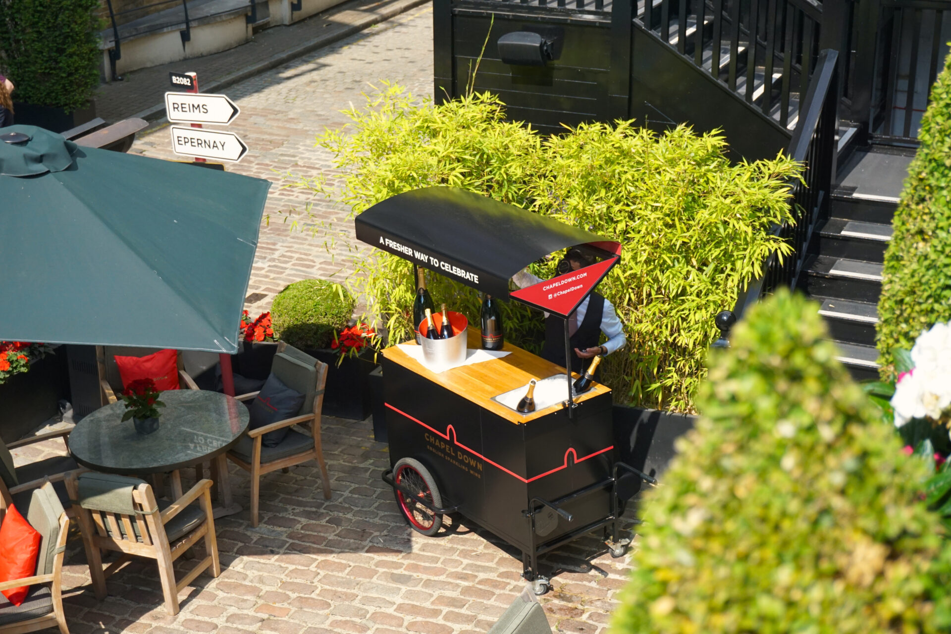 A black champagne cart with bottles in ice stands on a cobblestone patio near The American Bar, surrounded by green plants, outdoor seating with grey tables and chairs, and directional signs for Reims and Epernay.