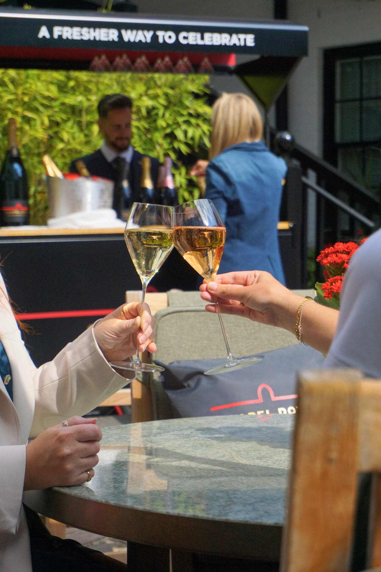 Two people clink wine glasses at an outdoor table, celebrating. The American Bar with bottles and an ice bucket is in the background, while two others stand nearby. The scene is bright and festive.