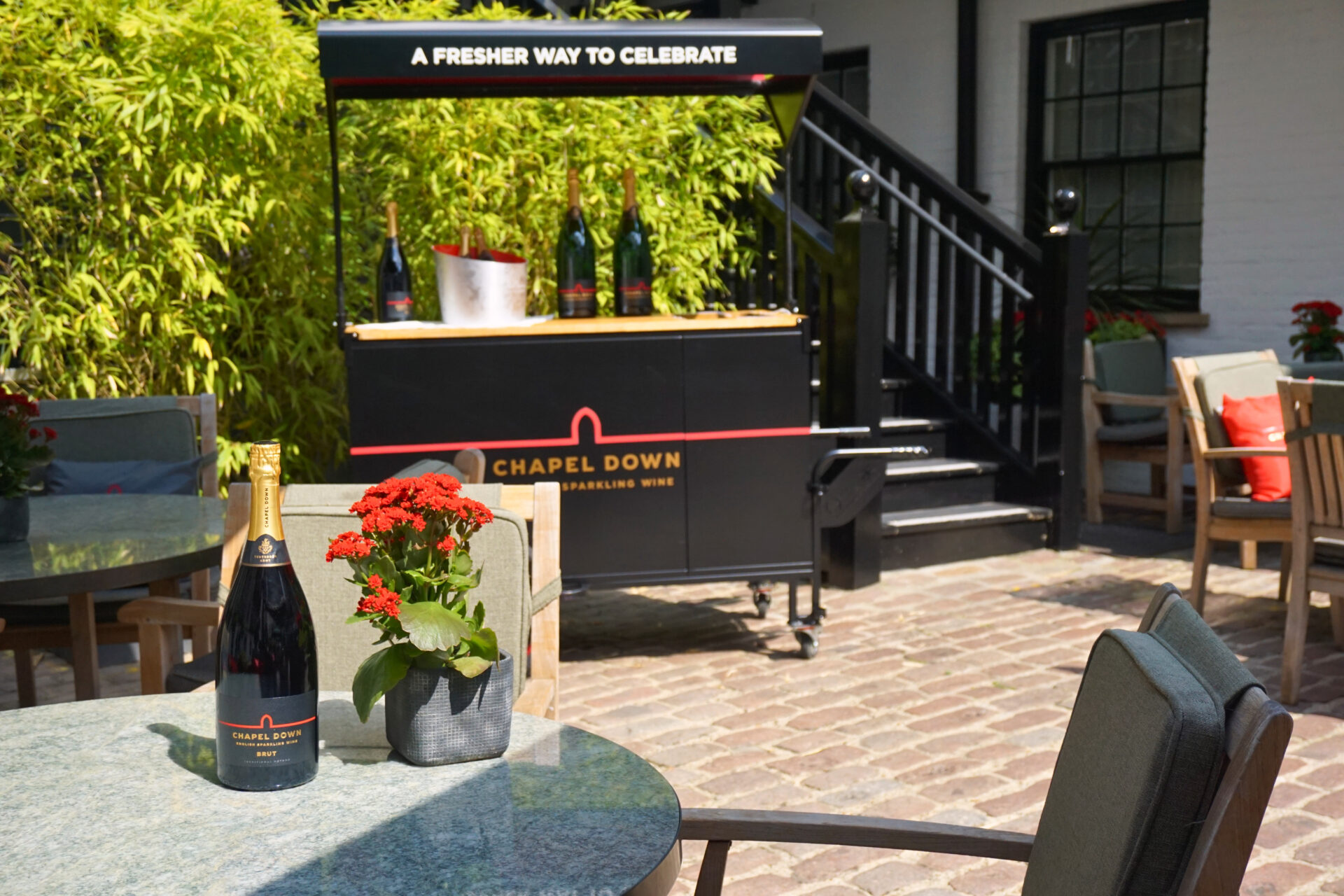 A bottle of Chapel Down sparkling wine and a potted red flower sit on a round outdoor table at The American Bar; in the background, a wine cart with bottles is surrounded by lush plants and inviting outdoor seating.