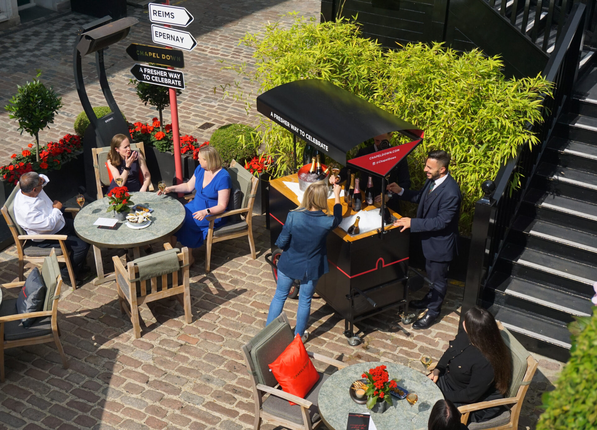 People sit at round tables in a sunny outdoor courtyard with flowers and greenery. A man from The American Bar serves drinks from a black cart while others relax, chat, and enjoy the atmosphere. Directional signs and black stairs are visible nearby.