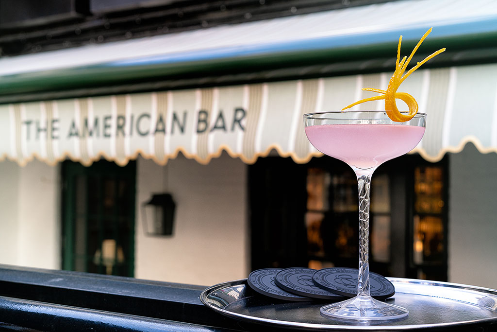 A pink cocktail in a coupe glass garnished with a twisted orange peel sits on a tray with coasters, set against The American Bar and the inviting ambiance of The Park Suite Terrace.