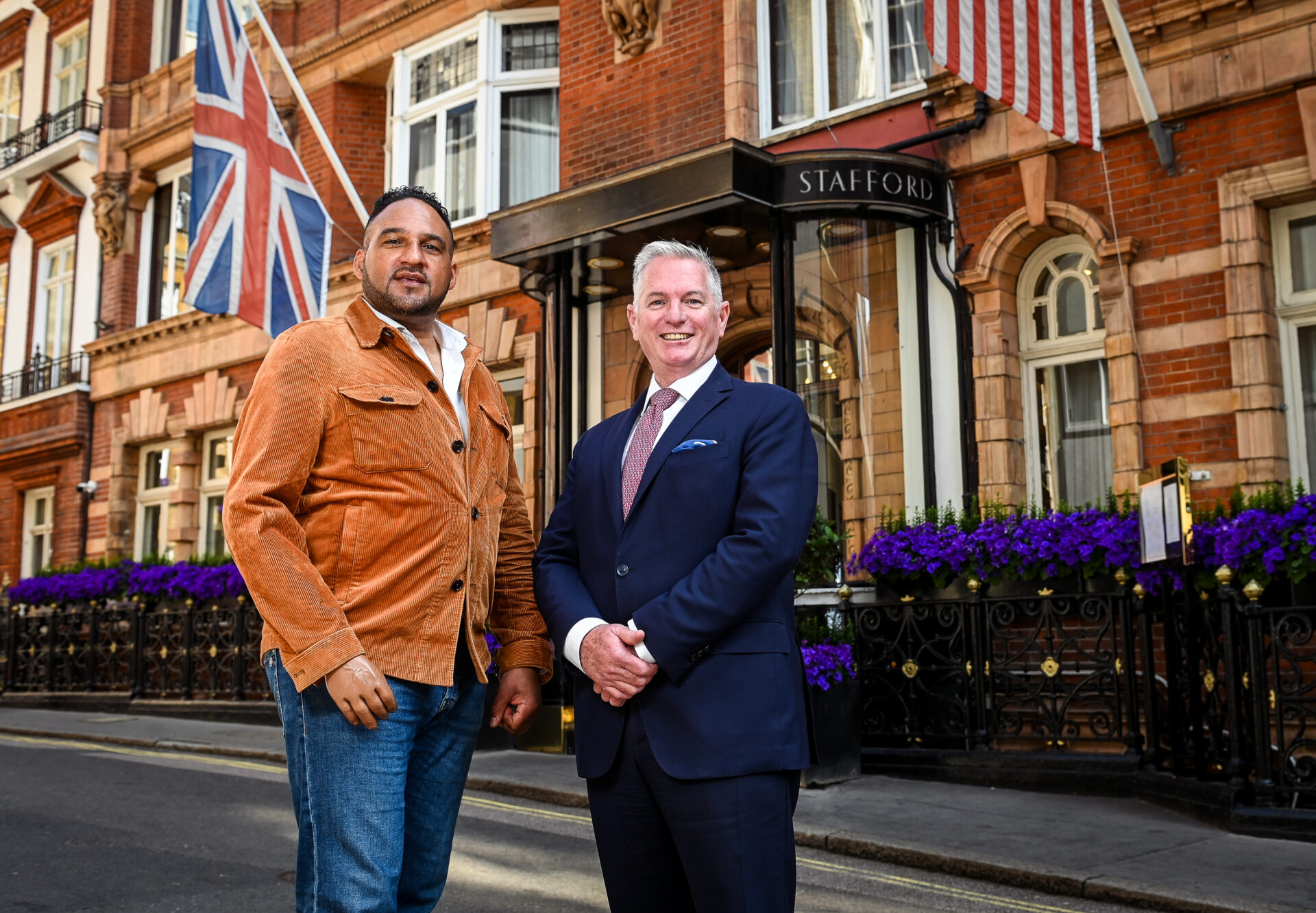 Two men, including Michael Caines, stand smiling outside The Stafford hotel in London; one wears a tan jacket and jeans, the other a navy suit. Behind them are British and US flags and a building with red brick and purple flowers.