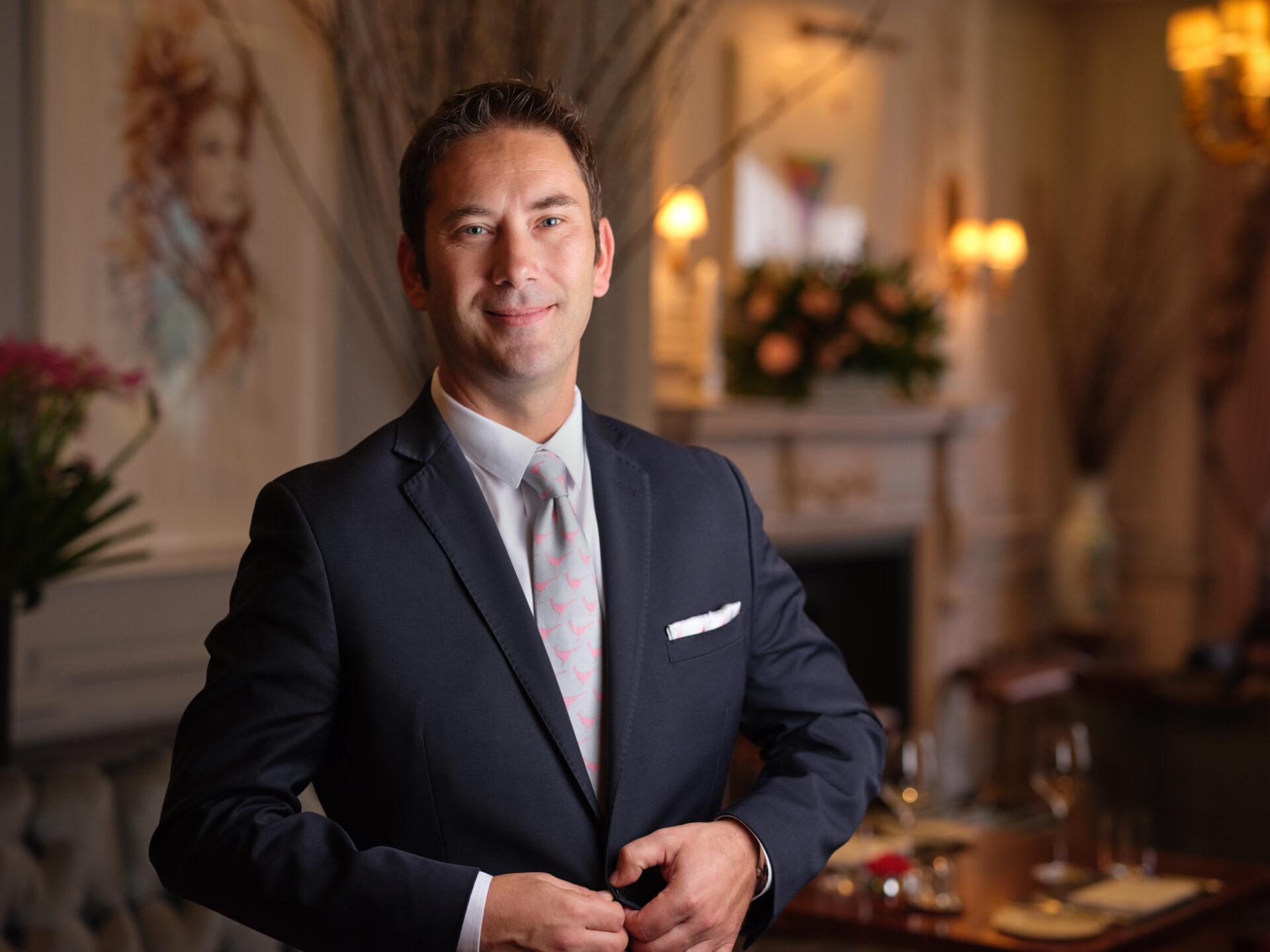 A man in a dark suit and light tie, reminiscent of Michael Caines sophisticated style, stands confidently in an elegant, softly lit dining room with floral decorations and art on the walls. He is smiling and buttoning his jacket.