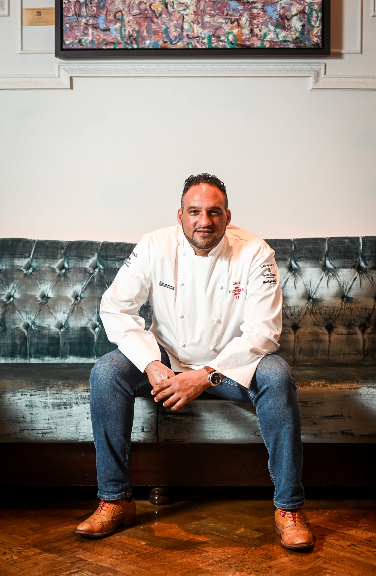 A chef in a white uniform and jeans, resembling Michael Caines, sits on a tufted teal sofa, hands resting on his knees, with a colorful painting hanging on the wall behind him.