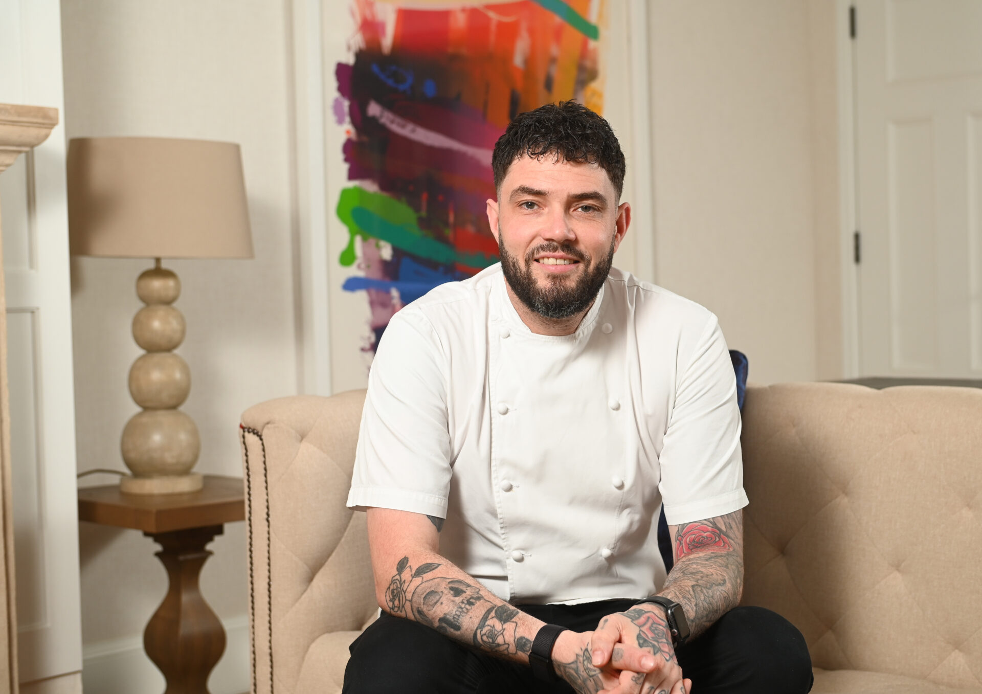A man with tattoos, wearing a white chef’s jacket, sits smiling on a beige sofa. Behind him is a colorful abstract painting and a lamp on a side table, evoking the relaxed elegance of Michael Caines’ signature style.