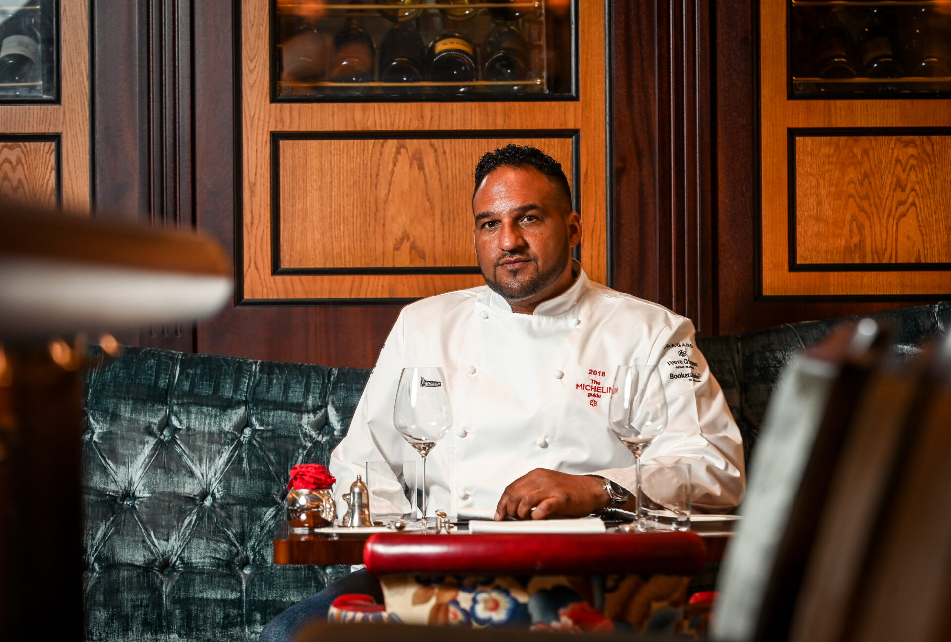 A chef in a white uniform sits at an elegant restaurant table set with empty wine glasses and cutlery, with dark wood paneling and wine bottles displayed behind him.