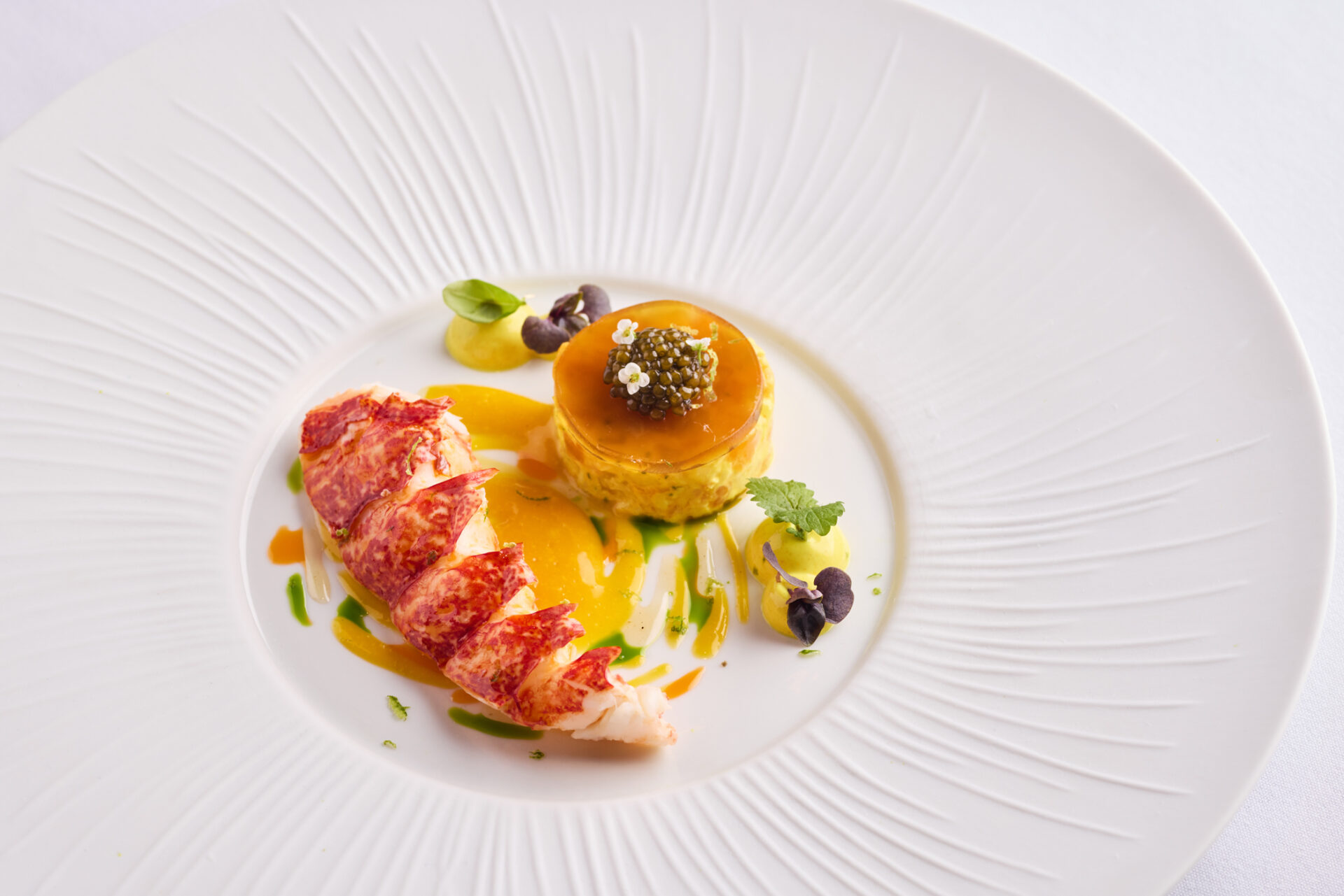 A gourmet dish at The Stafford London featuring lobster tail slices, a round savory mousse topped with caviar, and decorative sauces with microgreens on a white textured plate.