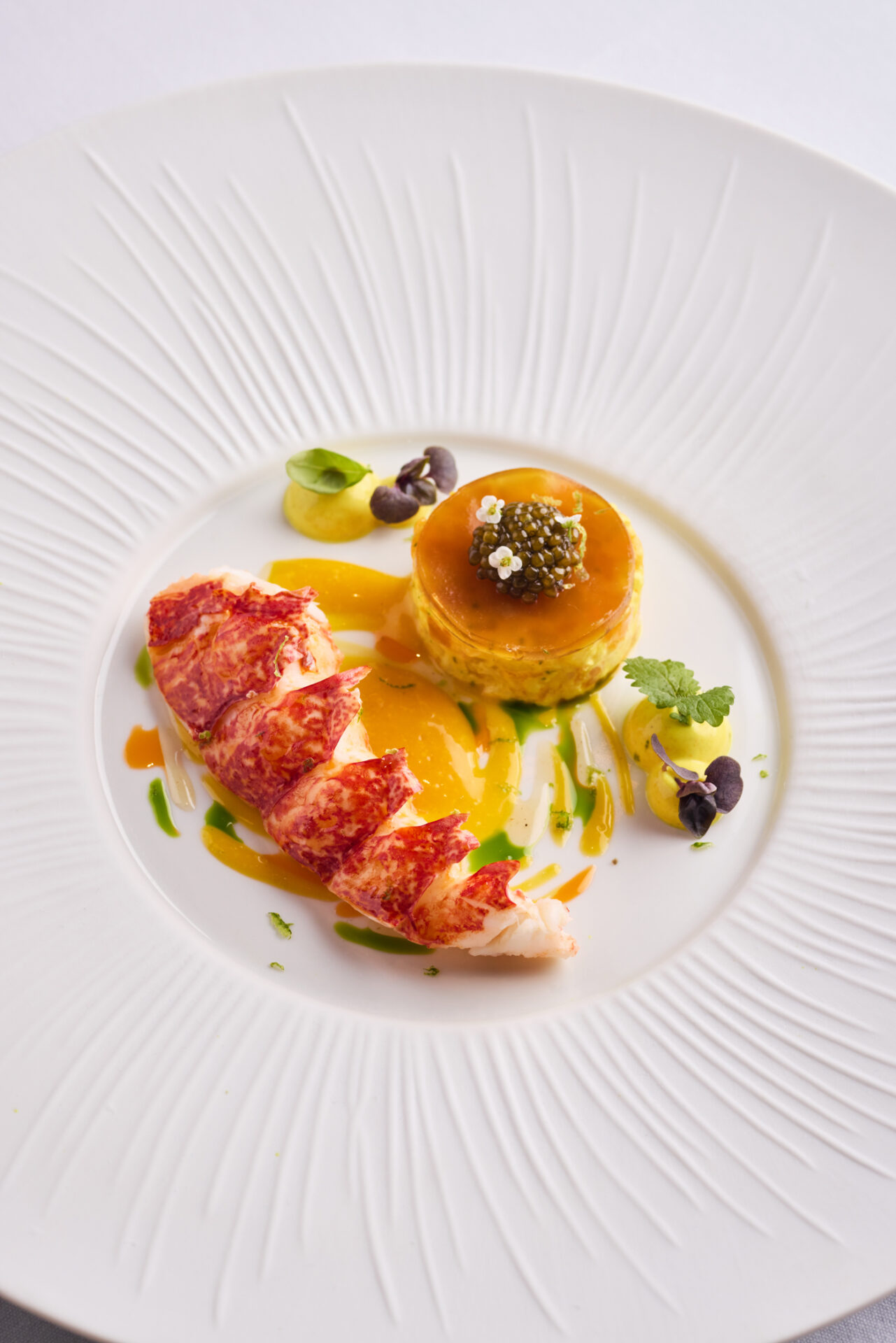 Sliced lobster tail next to a round custard topped with caviar and herbs, artfully plated with colorful sauce drizzles and microgreens on a white textured plate—an elegant highlight from The Game Bird menus.