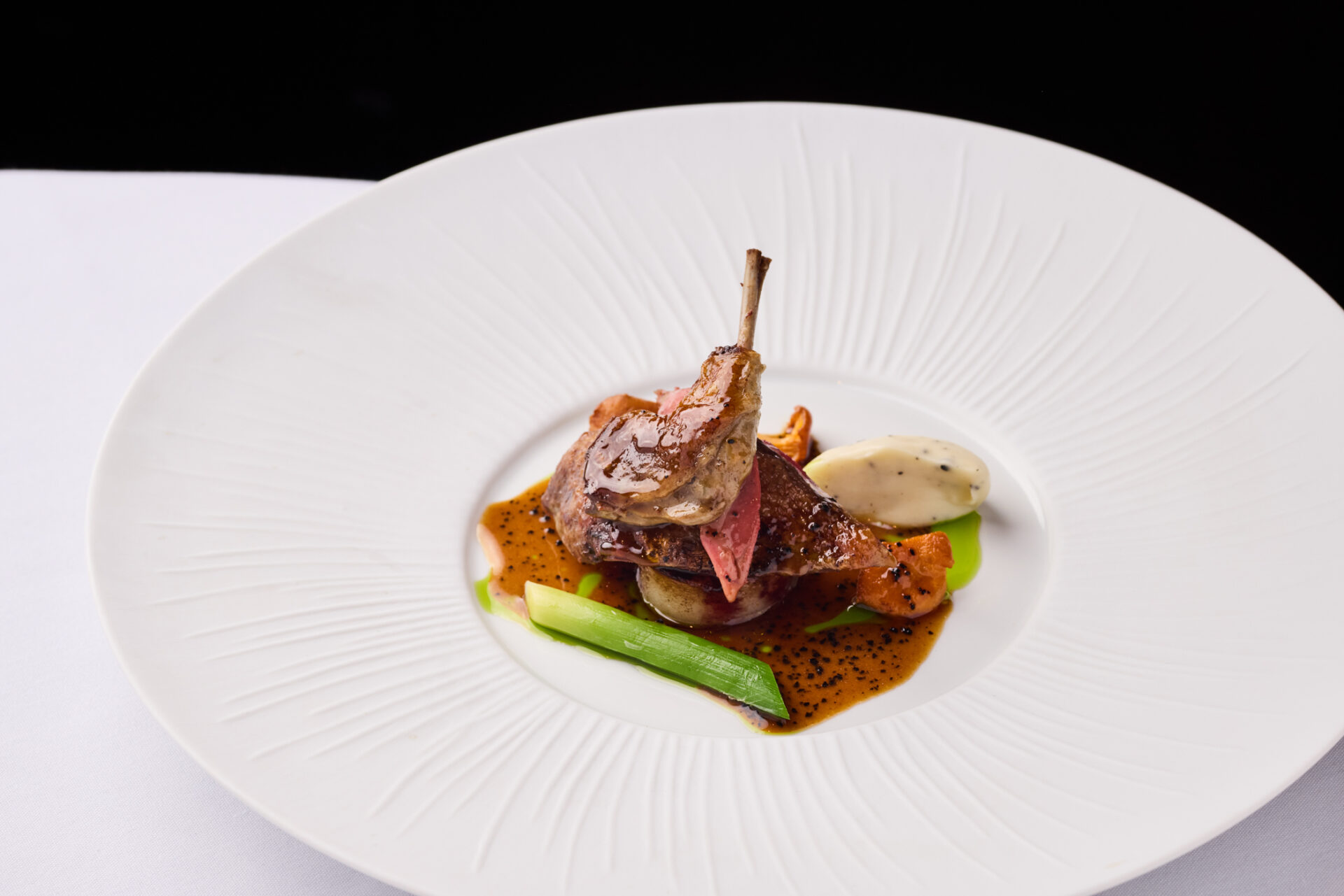 A gourmet dish from Michael Caines at The Stafford, featuring a roasted quail leg on a bed of vegetables, drizzled with rich brown sauce and finished with a dollop of creamy white sauce on an elegant white plate.
