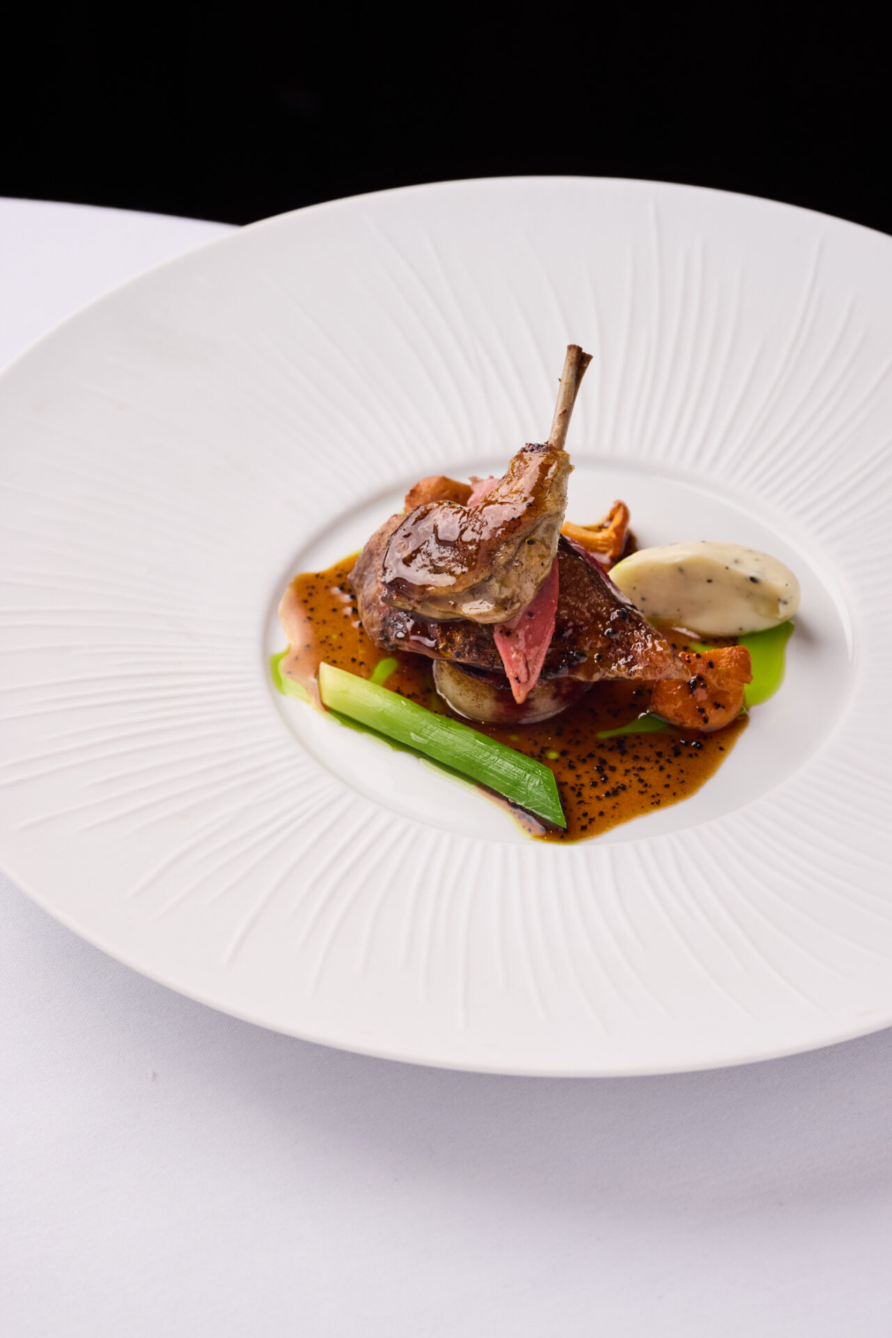 A gourmet dish featuring a seared piece of meat with a small bone, served with brown sauce, green vegetables, and a dollop of creamy white purée on an elegant white plate—perfect for enjoying exclusive Dining Offers.